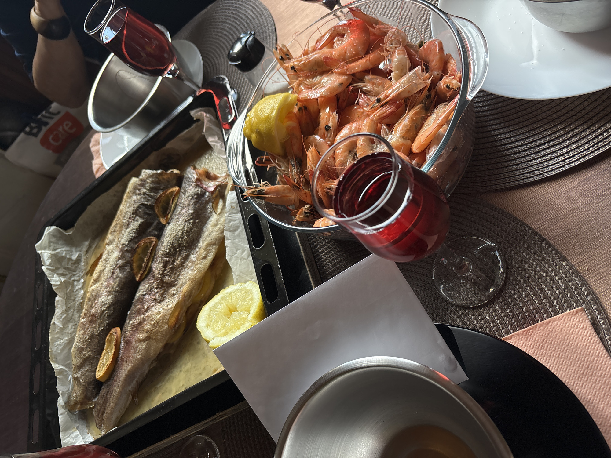 The image depicts a table setting with food and drinks. A clear glass bowl is filled with cooked shrimp and a lemon wedge. Next to it is a baking sheet lined with parchment paper, holding two cooked fish fillets garnished with lemon slices. A glass of red wine stands near the bowl of shrimp. Other objects include plates, a napkin, and cooking utensils. The setting suggests a meal is being prepared or enjoyed. It is impossible to determine if this scene is located in Varna, Bulgaria, or any other specific location.
