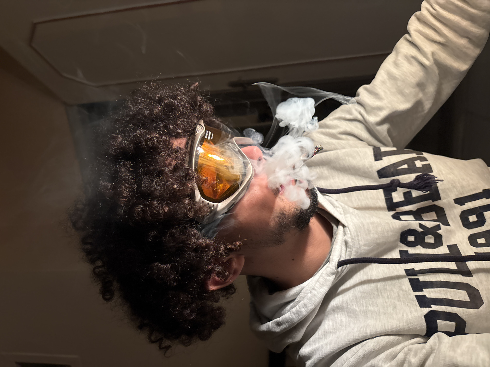 A man with curly hair, facial hair, and wearing orange-tinted goggles is exhaling smoke. He is wearing a grey hoodie with black lettering. The location is likely indoors. The city of Tanger, Morocco, cannot be confirmed from the image.