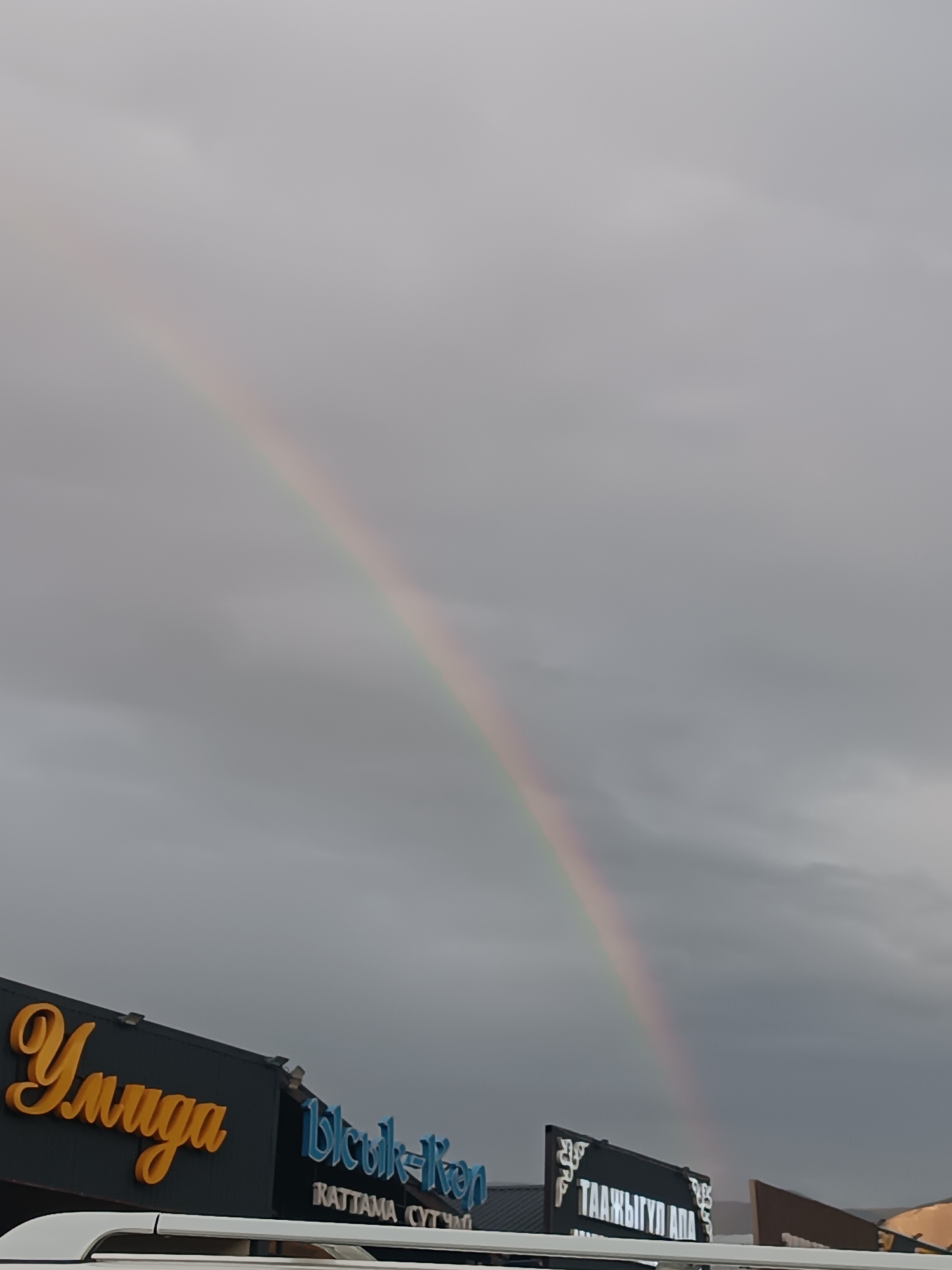 In the city of Sasyk-Bulak, Kyrgyzstan, a rainbow arcs across a cloudy sky, its colors faintly visible against the gray. Below, the facades of buildings display signage, including the word "Уииigа" in large, yellow, stylized lettering on a dark background, and another sign with blue lettering that appears to read "Ысык-Көл". Further to the right, a black sign with white decorative elements and text in an unfamiliar script, possibly Cyrillic, is partially visible, with the visible text appearing to be "ТААЖЫГҰП АПА". A white vehicle is positioned in the foreground, with its roofline partially obscuring the lower part of the building signs. The overall scene suggests a slightly overcast day with a fleeting natural phenomenon.