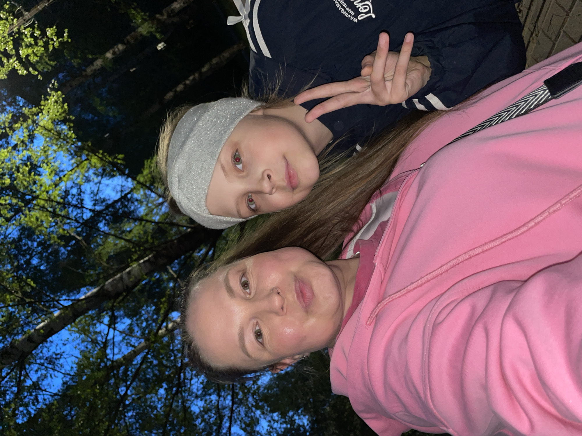 Two people are posing for a photo, likely a selfie. One person is an adult wearing a pink hoodie and the other is a younger person wearing a dark jacket with white stripes and a silver headband. The younger person is holding up two fingers in a peace sign. The photo is taken outdoors, possibly in a park or wooded area, as trees and blue sky are visible in the background. The image is taken at night or in low light conditions. The location is Izhevsk, Russia.