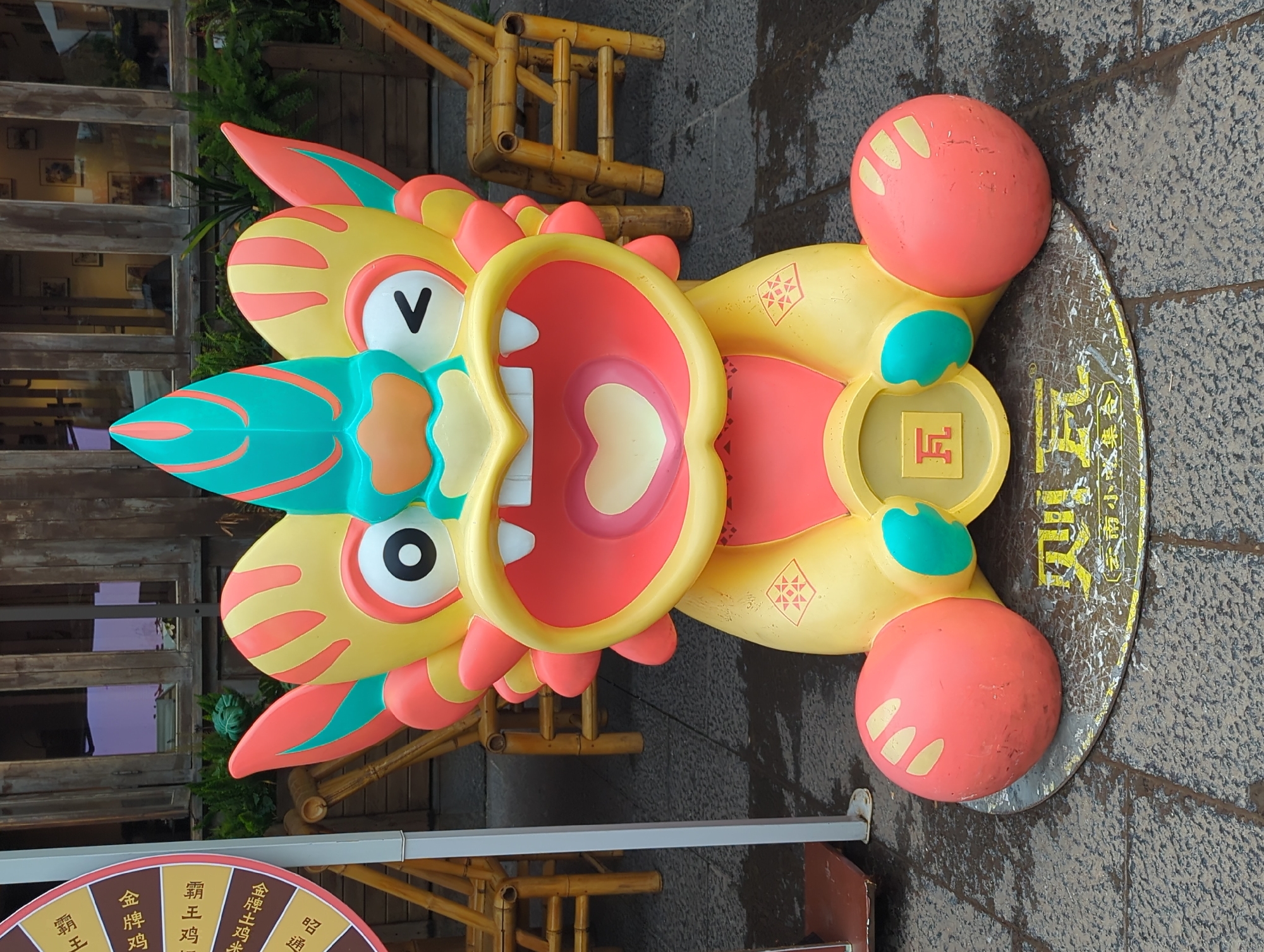This is a vertical, eye-level, full shot of a large, colorful, cartoon-like sculpture of a mythical creature, possibly a dragon or phoenix, positioned outdoors on a stone-tiled surface. The sculpture is primarily yellow with accents of orange, red, and teal. It features a large, open mouth with stylized teeth, wide eyes with black pupils, and decorative elements resembling flames or feathers. Two large, round, orange feet are visible on either side of the sculpture, and a circular golden emblem with Chinese characters is attached to its chest. The creature appears to be smiling or roaring.
In the background, there is a wooden structure with bamboo elements, suggesting an outdoor market or a decorative display. Some greenery is also visible behind the sculpture. A portion of a spinning wheel with Chinese characters, possibly a game or prize wheel, is visible in the lower left corner of the image. The ground is wet, indicating recent rain or damp conditions. The overall impression is that of a festive or promotional display, likely in a public area.
Visible text in the image includes:
- "V" on the creature's face.
- A stylized "F" on the golden emblem.
- Chinese characters on the golden emblem and the spinning wheel, including "云南小吃集合" (Yunnan Snack Collection), "昭通" (Zhaotong), "金牌鸡" (Golden Chicken), and "霸王鸡" (Overlord Chicken).