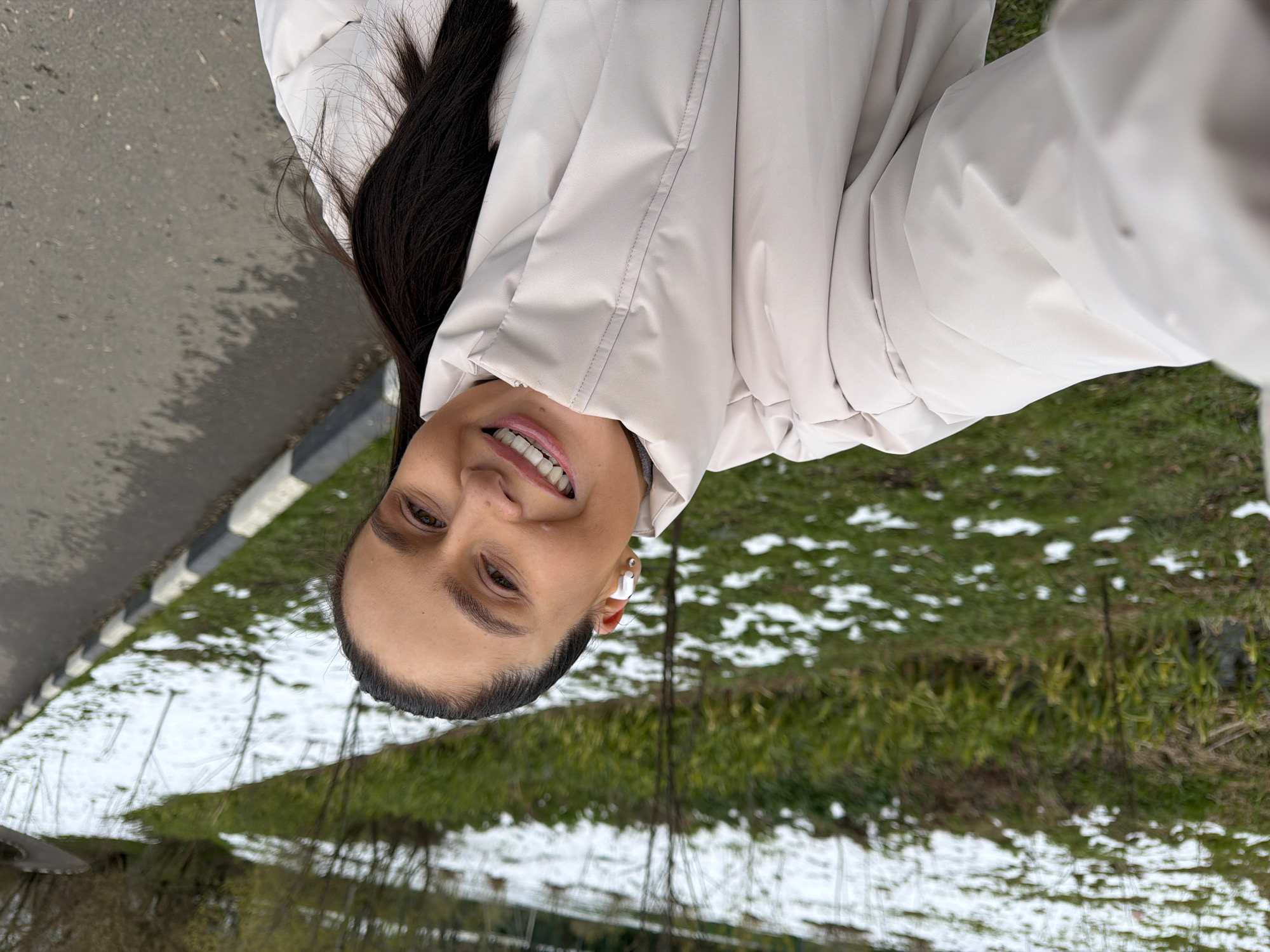 An outdoor portrait captures a smiling woman looking directly at the camera, likely taking a selfie. She has dark hair, pulled back, and is wearing a light beige or off-white puffer jacket. A white wireless earbud is visible in her right ear, and she displays a bright smile.
The setting appears to be a park or roadside area in Khimki, Russia, during late winter or early spring. To the woman's left is a paved path or road, bordered by a black and white striped curb. The ground to her right and in the background consists of green grass dotted with patches of melting snow. Bare trees and shrubs are visible in the middle distance. The soft, even lighting suggests an overcast daytime sky. The woman's expression conveys happiness or contentment. There is no visible text in the image.
