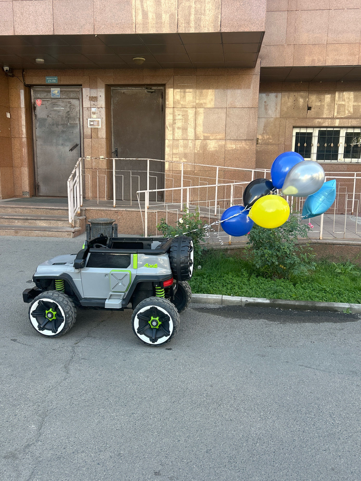 A grey toy Jeep with green accents and large wheels is parked on an asphalt surface in front of a building. The Jeep has "4x4" written on its side in green. A bunch of balloons, including blue, black, yellow, and silver, are tied to the Jeep and float upwards. The building behind the Jeep has brown tiled exterior walls, two metal doors, and a window with bars. A metal railing and stairs lead up to one of the doors. Green grass and some plants are visible in front of the building. The scene appears to be in an urban environment, possibly an apartment complex entrance in Almaty, Kazakhstan, indicated by the signage on the door.