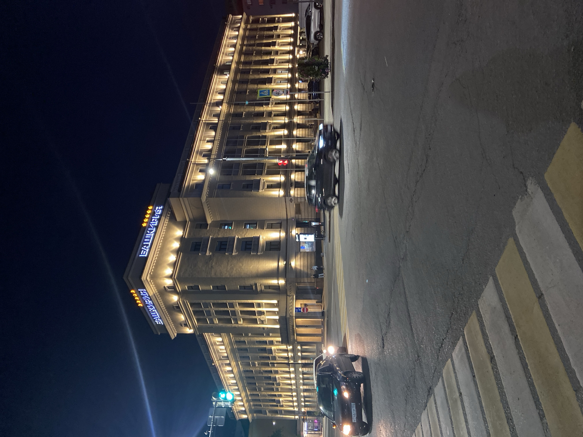 The image depicts a night scene in Ufa, Russia, focusing on a grand, illuminated building with signage that reads "БАШКИРИЯ" in neon blue lights, indicating it is likely a hotel or a prominent establishment. The building is multi-storied with many windows, each lit from within, creating a warm and inviting glow against the dark night sky. Yellow lights line the exterior of the building, highlighting its architectural details.
In the foreground, a street with a pedestrian crossing is visible. Cars are driving on the road, with one black car in the immediate foreground, its headlights illuminating the pavement and a portion of the pedestrian crossing. The car has a license plate visible, though the text is partially obscured. Another car is further down the street, also illuminated.
The scene appears to be urban, with streetlights and traffic signals also present, including a red traffic light. The overall atmosphere is one of a bustling city at night, with the illuminated building serving as a central focal point. The weather appears clear, given the distinct visibility of the night sky and the building's lights. The image is shot with a slight tilt, adding a dynamic perspective to the scene.