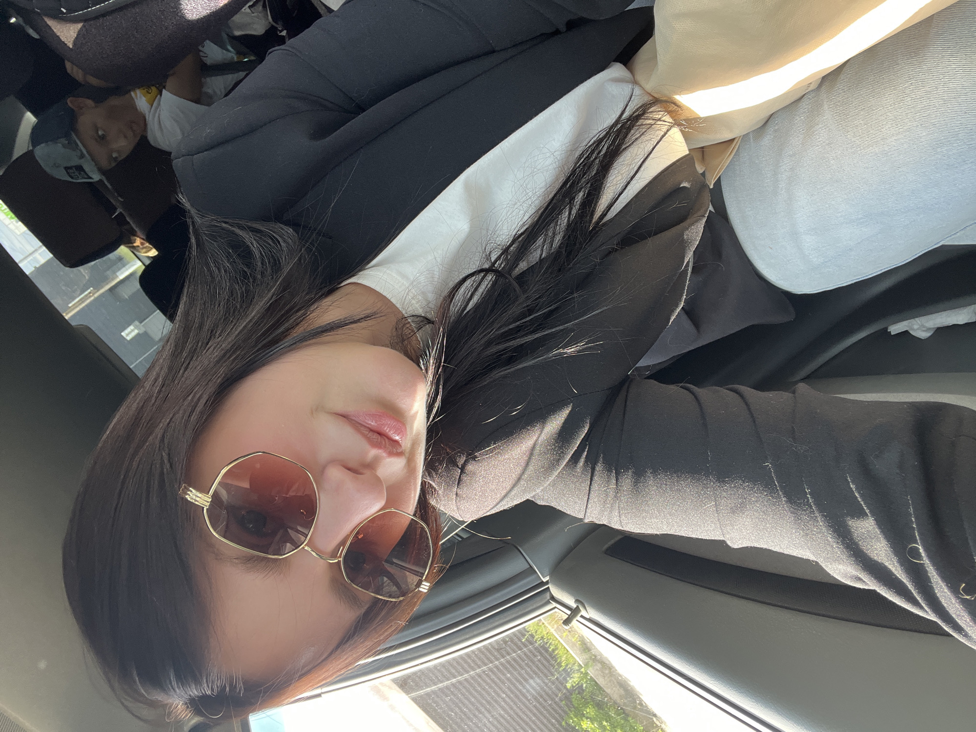 A woman is taking a selfie in a car. She has long, dark hair and is wearing sunglasses with gold frames, a white t-shirt, and a black blazer. She is smiling. In the background, a young boy wearing a baseball cap is visible in the back seat. The car appears to be in motion, as indicated by the sunlight streaming through the window and the blurred background. The image does not contain any specific visual cues that would allow for identification of the location within the city of Chaikovsky, Russia.