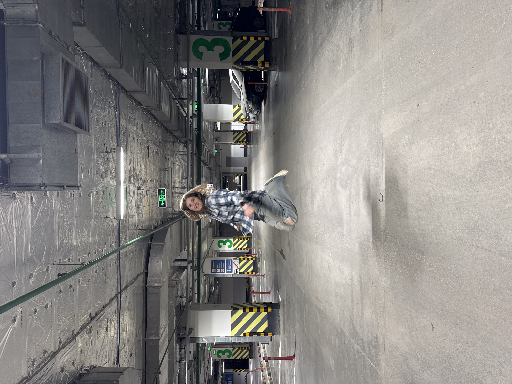 The image depicts an underground parking garage in Perm, Russia, with a young woman captured mid-jump in the foreground. The woman has light brown hair and is wearing a blue and white plaid shirt, ripped blue jeans, and light-colored sneakers. She is smiling and appears joyful.
The parking garage features concrete flooring and ceiling, with the walls predominantly covered in a silver, reflective insulation material, possibly for sound or temperature control. Numerous metallic ventilation ducts run along the ceiling and walls. Overhead fluorescent lights illuminate the space.
In the background and mid-ground, several parked cars are visible, partially obscured by concrete pillars. The parking spots are marked by yellow and black striped barriers and red-topped safety posts. Green signs displaying the large number "3" are prominent on the pillars, indicating a specific level or section of the garage. Multiple green "EXIT" signs with arrows pointing to the left are also seen. Other informational signs include a "no stopping" symbol (red circle with a diagonal line over a car and person) and a white "P" with an arrow, indicating parking directions. The overall lighting is artificial and functional, typical of an indoor parking facility.