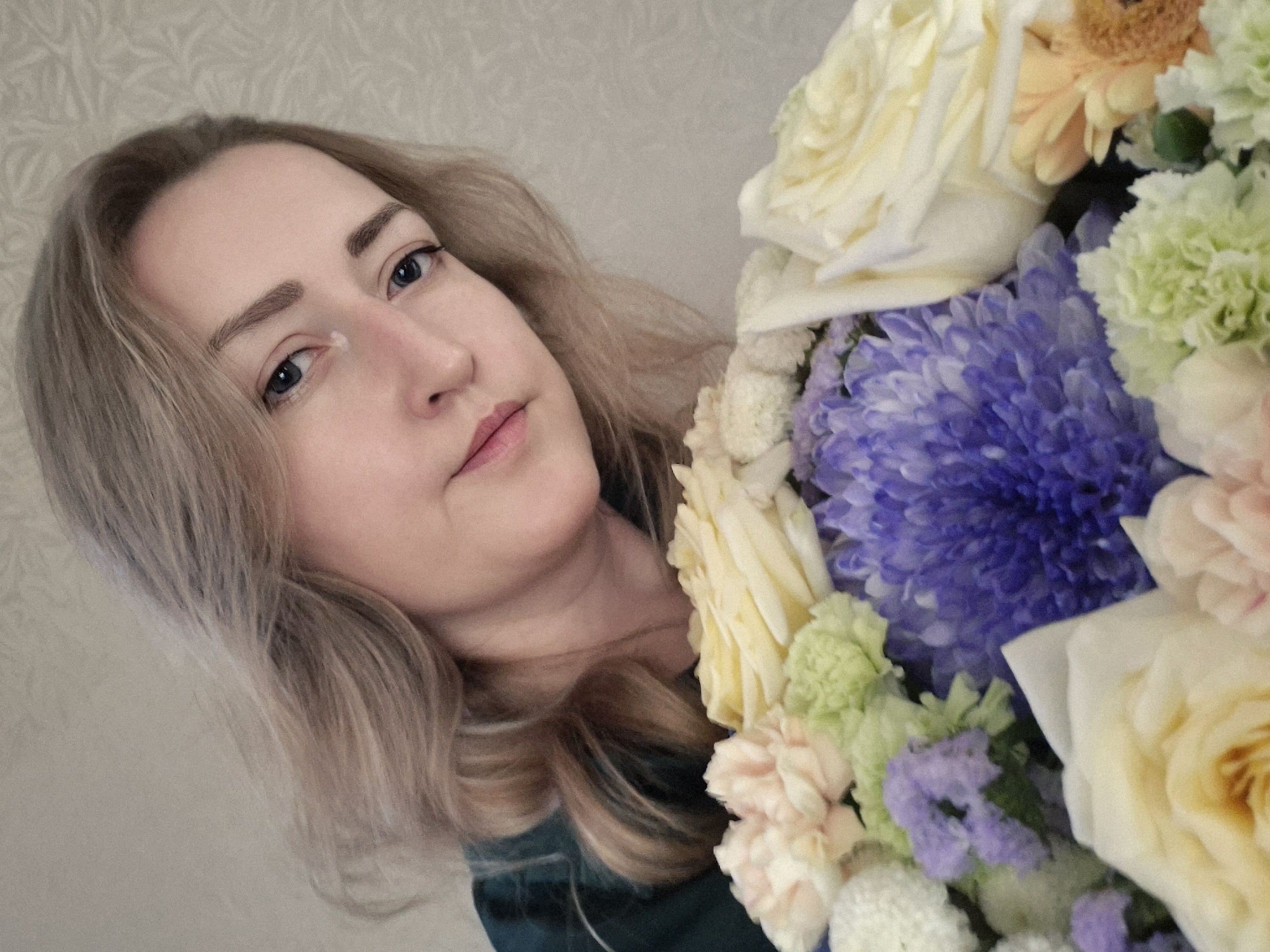 A medium shot captures a young woman holding a bouquet of flowers. She is looking towards the camera with a neutral expression. Her blonde hair is styled loosely, framing her face. The bouquet is composed of various flowers in shades of white, yellow, light pink, light green, and purple, with a prominent large purple chrysanthemum in the center. The background appears to be a textured wall. The image has a soft focus, giving it a dreamlike quality. The scene is indoors, and the lighting is soft. There is no indication of the specific location in Chaikovsky, Russia.