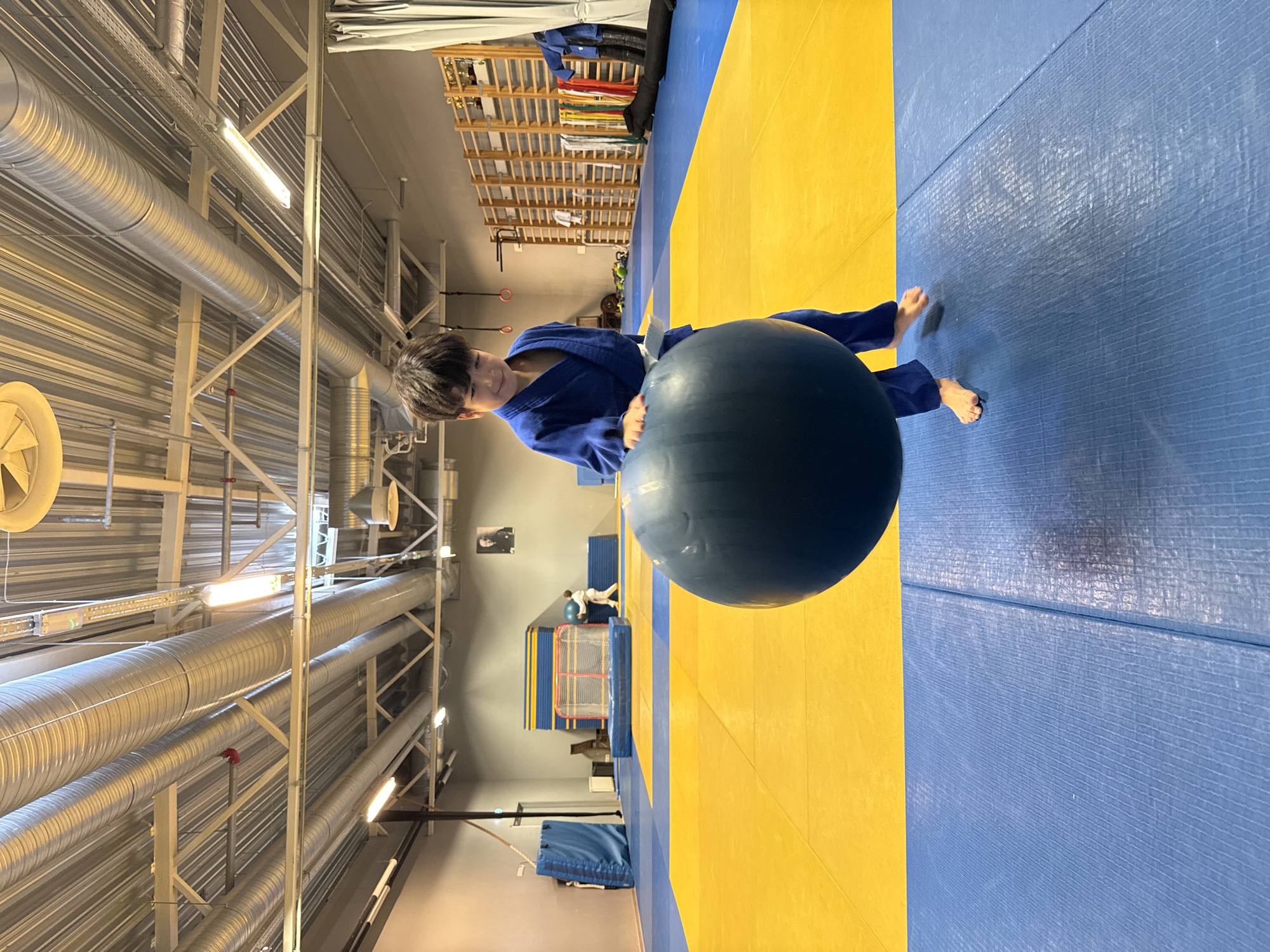 A young boy wearing a blue judo uniform with a white belt is holding a large, dark blue exercise ball. He is standing on a blue and yellow mat in what appears to be a dojo or a sports training facility. The room has a high ceiling with exposed metal ducts and lighting fixtures. In the background, there are various sports equipment, including mats, a hockey net, and shelves with colorful belts. There is a framed picture on the wall. The boy is looking down at the ball with a slight smile, and his left foot is slightly raised. The image is captured from a low angle, focusing on the boy and the ball. This scene is likely in Tallinn, Estonia.