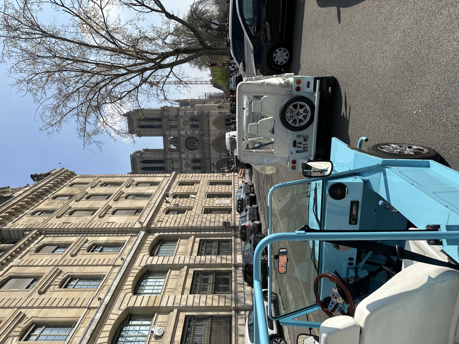 The scene captures an urban street in Paris, France, with a direct view of Notre Dame Cathedral. In the immediate foreground, the interior and front-right exterior of a light blue, open-top, two-door Moke-style vehicle are visible, featuring white seats, a brown wooden steering wheel, and a water bottle in the passenger footwell. Directly ahead, a white, open-top Moke-style vehicle is positioned, displaying a white fabric roof, white seats, and a spare tire mounted on its rear, with a French license plate reading "FA-991-MS".
To the left, a multi-story building constructed of light-colored stone with numerous windows, some fitted with closed shutters, dominates the streetscape. Multiple parked and moving passenger vehicles, including dark-colored sedans and hatchbacks, occupy the street in the midground. A large, bare deciduous tree with intricate branches stands on the right side of the midground.
In the background, the western facade of Notre Dame Cathedral is clearly visible, featuring its two iconic towers and central rose window. Extensive scaffolding and a large construction crane are present behind and to the right of the cathedral, indicating ongoing restoration work. Indistinct figures of people are visible in the far background near the cathedral and a traffic light on the right. The sky is clear and blue, suggesting a sunny day.