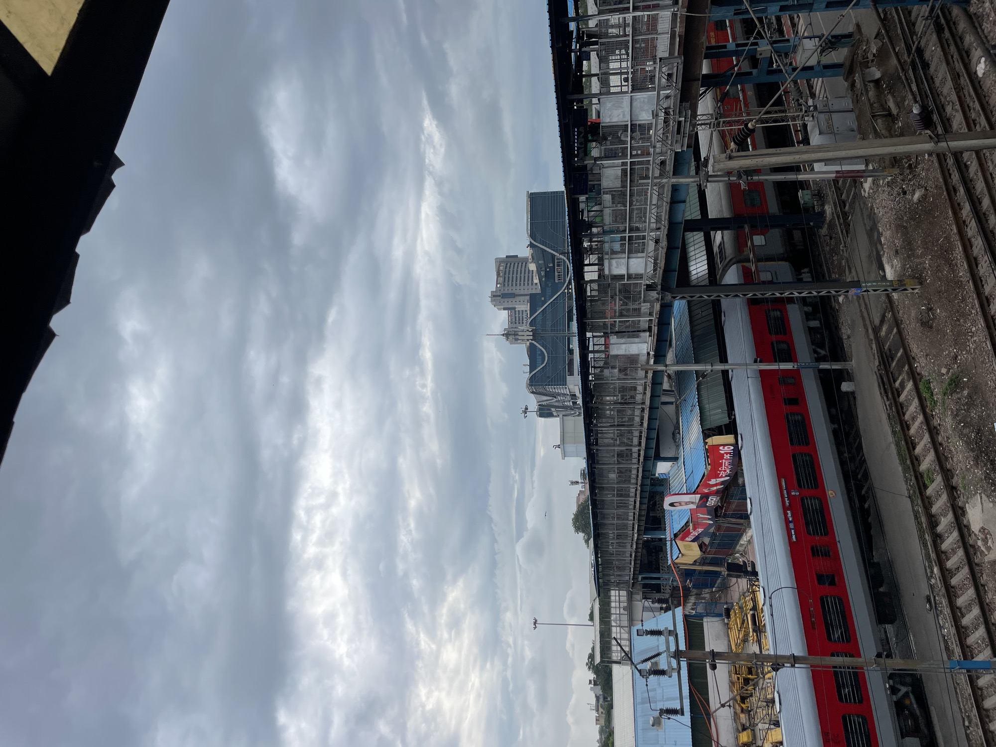 The image shows a view of a railway station in New Delhi, India, on a cloudy day. In the foreground, a red and gray train is on the tracks. Above the train, a pedestrian overpass with glass walls and a metal roof spans across the tracks. In the background, there are several buildings, including a large, modern office building with a distinctive curved facade. The sky is overcast with heavy clouds. The scene depicts a typical urban railway station environment, with infrastructure for transportation and commercial activity.