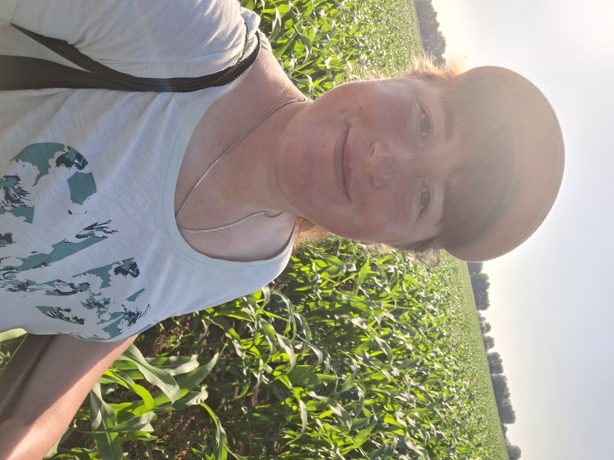 A woman wearing a light blue t-shirt, a brown baseball cap, and a silver necklace takes a selfie in a cornfield. The sun is bright and behind her, causing a lens flare. The corn stalks are tall and green, and the sky is clear and blue. The woman is smiling at the camera. The location is Unknown, Unknown.