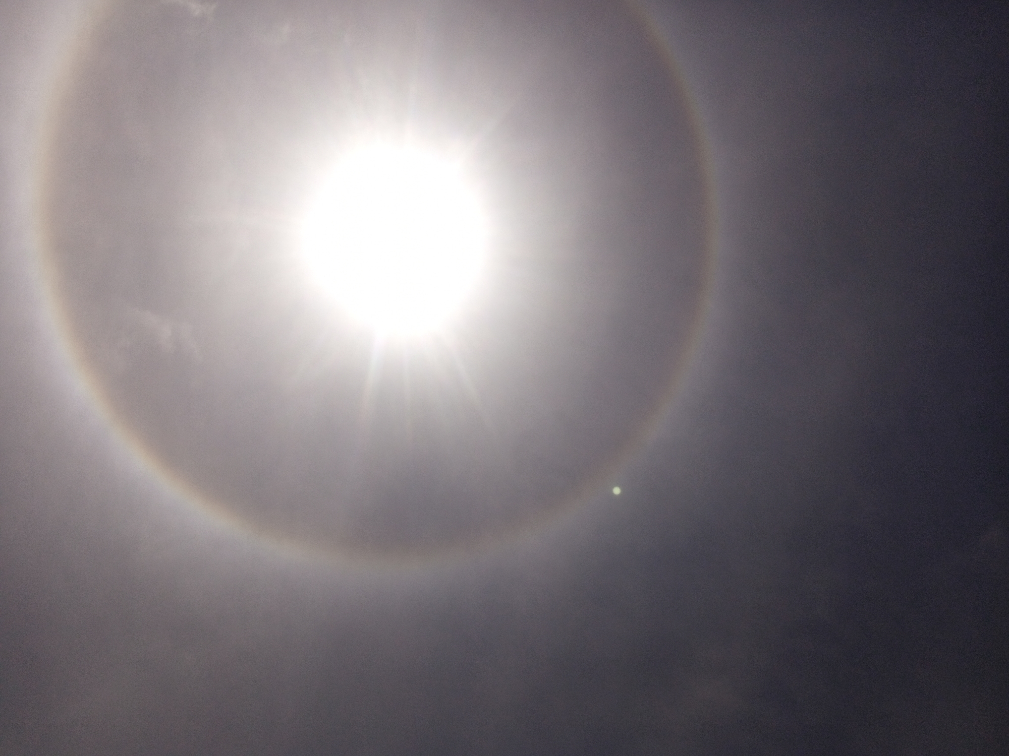This is a photograph of a sun halo, an optical phenomenon caused by sunlight refracting through ice crystals in the atmosphere. The sun is the brightest subject in the image, appearing as an intense white disc with rays emanating from it. A faint, circular halo surrounds the sun, with subtle color variations visible, suggesting a spectrum of light. The sky appears overcast and hazy, with a darker, textured atmosphere that is lighter and brighter near the sun. There are no people or man-made objects visible in the frame, nor any text. The image captures a natural atmospheric event, likely during daylight hours, given the presence of the sun. The overall mood is serene, dominated by the celestial display.