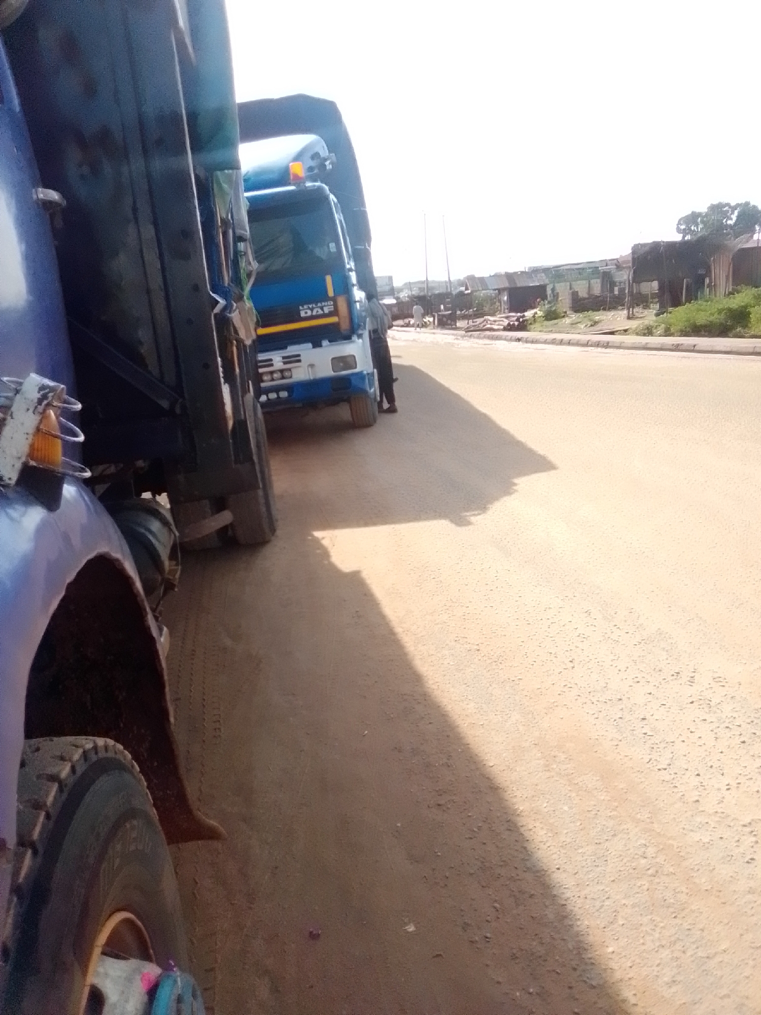 The image depicts a scene on a dirt road in Kwadon, Nigeria. In the foreground, the side of a blue truck is visible, with its large tire and fender on the left. Behind it, another blue truck with "LEYLAND DAF" lettering on its side is parked. The trucks are positioned on the right side of the road, casting shadows. The road itself is dusty and uneven. Along the left side of the road, a row of modest buildings and structures, some appearing to be informal shops or dwellings, are visible in the background. A few scattered trees and dense green foliage can also be seen. The sky is bright and hazy, suggesting a sunny day. There are a few individuals present in the scene, including one person standing near the front of the blue "LEYLAND DAF" truck and another figure walking further down the road. The overall impression is of a typical day in a developing urban or semi-urban area.