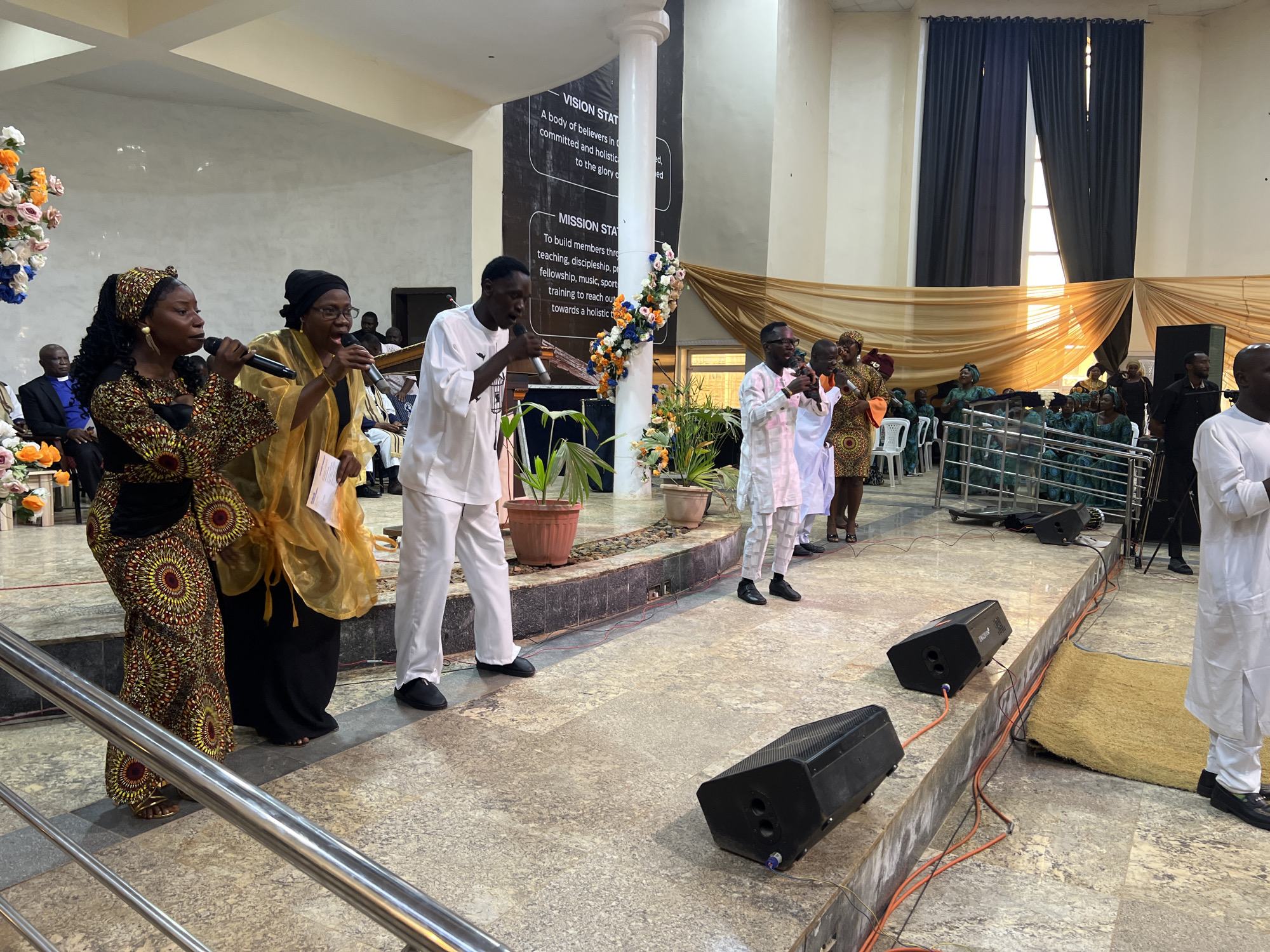The image depicts a religious gathering in a large indoor hall, possibly a church in Kabong, Nigeria. Several people are present, with a group of five individuals in the foreground holding microphones and appearing to be singing or speaking. One person in the foreground, a woman in a colorful patterned dress and headwrap, is holding a microphone and singing. To her right, another woman in a yellow garment and black headscarf is also holding a microphone. Two men in white traditional attire are also holding microphones, and another man in white is partially visible on the far right. Behind them, a large banner with text, possibly a vision or mission statement, is displayed on a pillar decorated with flowers. Other individuals are seated or standing in the background, some of whom appear to be part of a choir. The hall has a tiled floor and is decorated with draped fabric and potted plants. Several black speaker boxes are placed on the floor, connected by visible orange wires. The overall atmosphere appears to be one of a lively religious service or event.