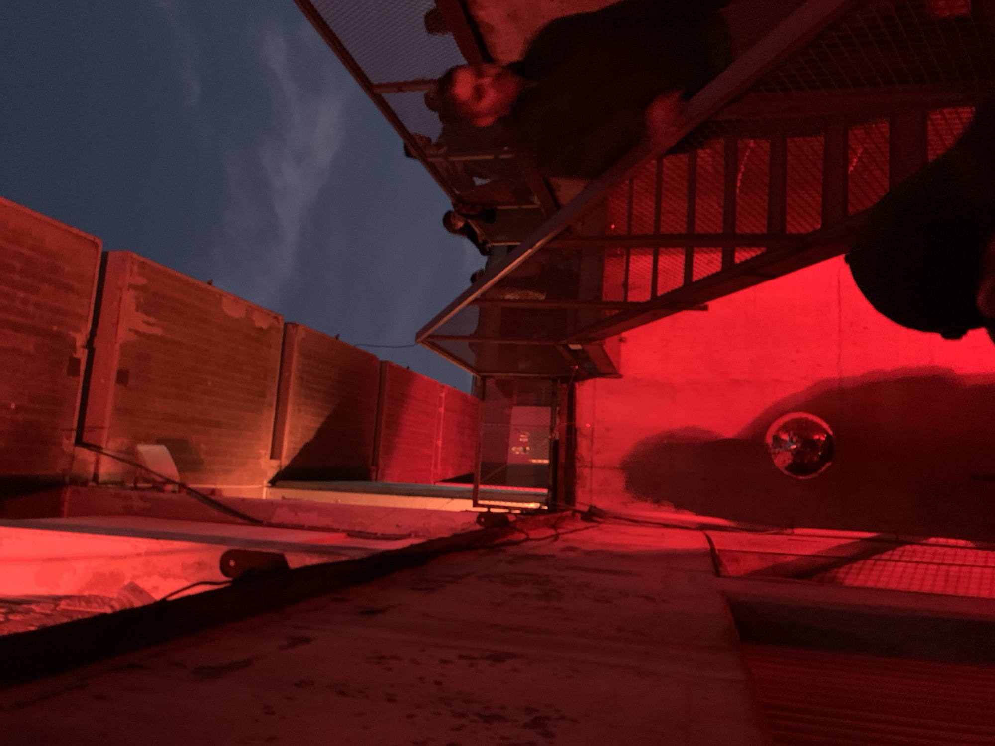The image depicts an urban industrial environment in Tel Aviv, Israel, likely situated beneath an elevated concrete structure such as a bridge or overpass. The scene is illuminated predominantly by intense red artificial light, suggesting dusk or night, with a dark blue sky and faint clouds visible in the upper right quadrant.
A multi-level metal staircase with mesh-like steps and metal railings ascends from the lower right towards the upper left. A small landing with a metal railing is visible midway up the stairs. To the left, a rough, textured concrete wall with visible wiring is strongly bathed in the red light. Overhead, large, heavy concrete beams form the underside of the elevated structure.
Approximately five to eight individuals are present. In the mid-ground, a person in a dark jacket is seen descending the stairs, partially blurred, with their face somewhat obscured. Further up the staircase, several other figures are visible, mostly in silhouette or blurred, either moving or standing. The top of a head wearing a dark cap is visible in the immediate lower foreground, centered. On the lower section of the red-lit concrete wall on the left, a small, circular, reflective object (resembling a fragment of a disco ball) is affixed, casting a distinct, tall, human-shaped shadow behind it. People are actively traversing the staircase, suggesting movement through a public or event-related space.
