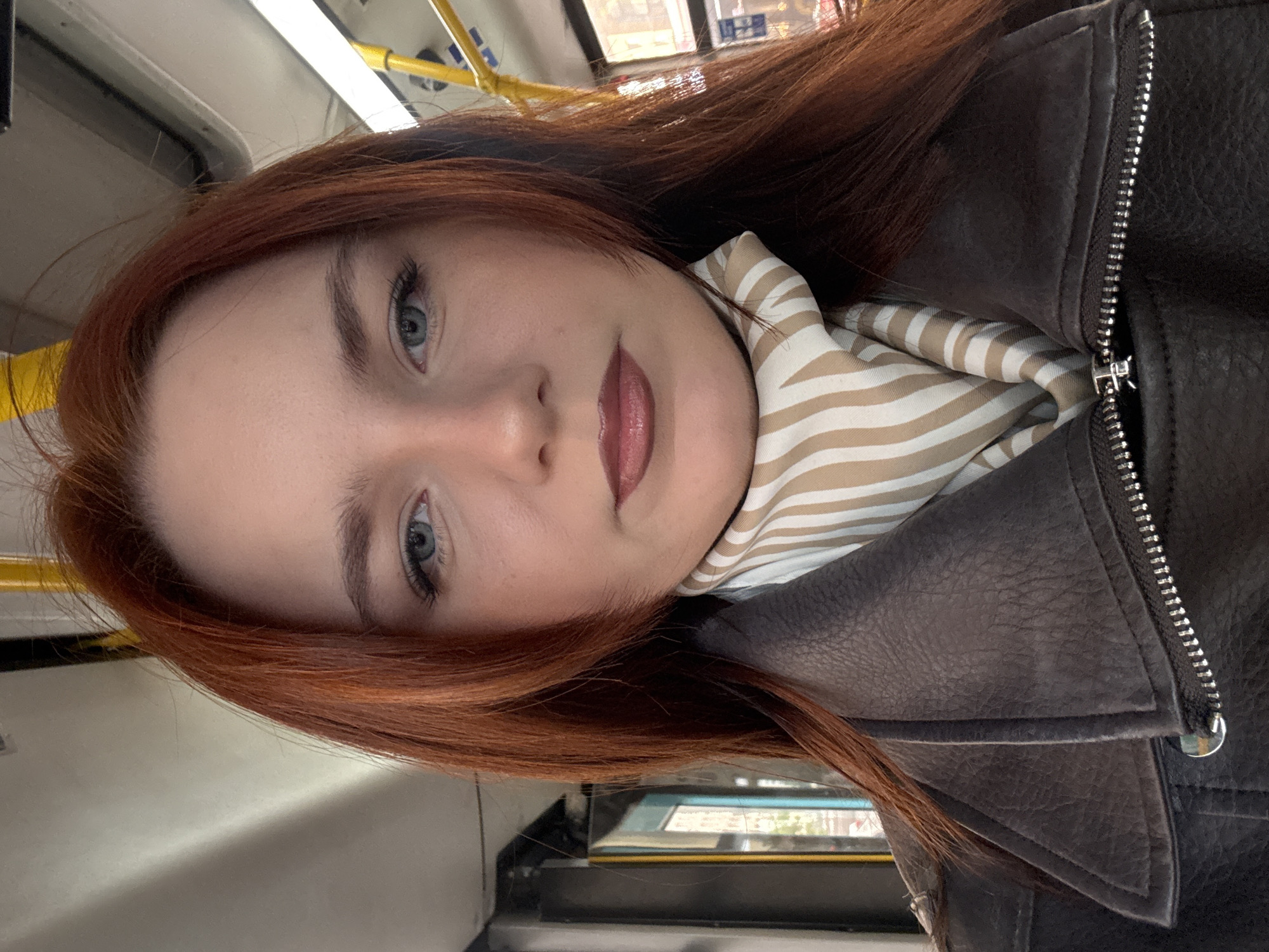 A selfie-style photograph of a woman with red hair and light eyes, taken in what appears to be the interior of a public transportation vehicle, potentially a bus or train. She wears a brown leather jacket and a scarf with a beige and white striped pattern. The background is blurry, but elements of the vehicle's interior, such as yellow handrails, are visible. The location is unlikely to be Sinyavino, Russia, given that public transportation interiors often differ significantly across regions. Furthermore, without additional visual or contextual clues specific to Sinyavino, it's impossible to confirm that location based solely on this image. The focus is on the woman's face, capturing details like her skin tone and eye color.