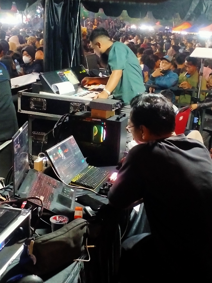 The image shows a man in a dark green shirt working on a mixing board in a dimly lit setting. He is facing right, interacting with the controls of the mixing board. He has dark hair and is wearing a beard. Another man, wearing a black shirt and glasses, sits behind him, facing left, and using a laptop. Both men appear to be working on sound and video equipment. The scene is in a large, crowded area with people gathering in the background. There are two black speaker cases to the left of the men. The background is a night scene, with lights from the crowd behind the men. Based on the provided coordinates, the scene appears to be in a public space in Boyolali, Indonesia.