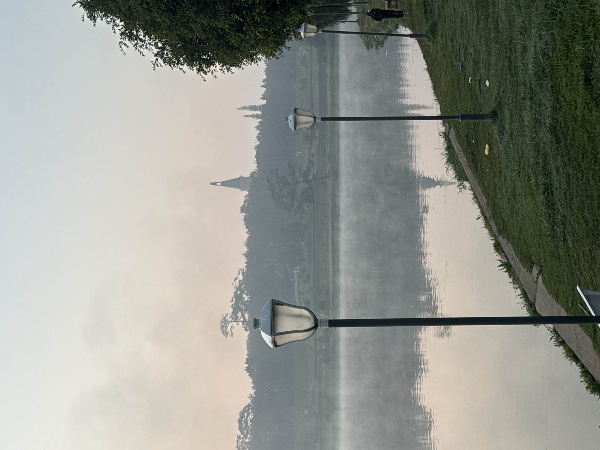 The image shows a misty morning view of Xuân Hương Lake in Đà Lạt, Vietnam. The lake reflects the surrounding trees and a distant spire. Several black lamp posts with white lamps are placed along the edge of the lake. The grass adjacent to the lake is green and slightly unkempt, with some litter scattered on it. A person is visible in the background walking along the edge of the lake. The sky is overcast, creating a hazy atmosphere.