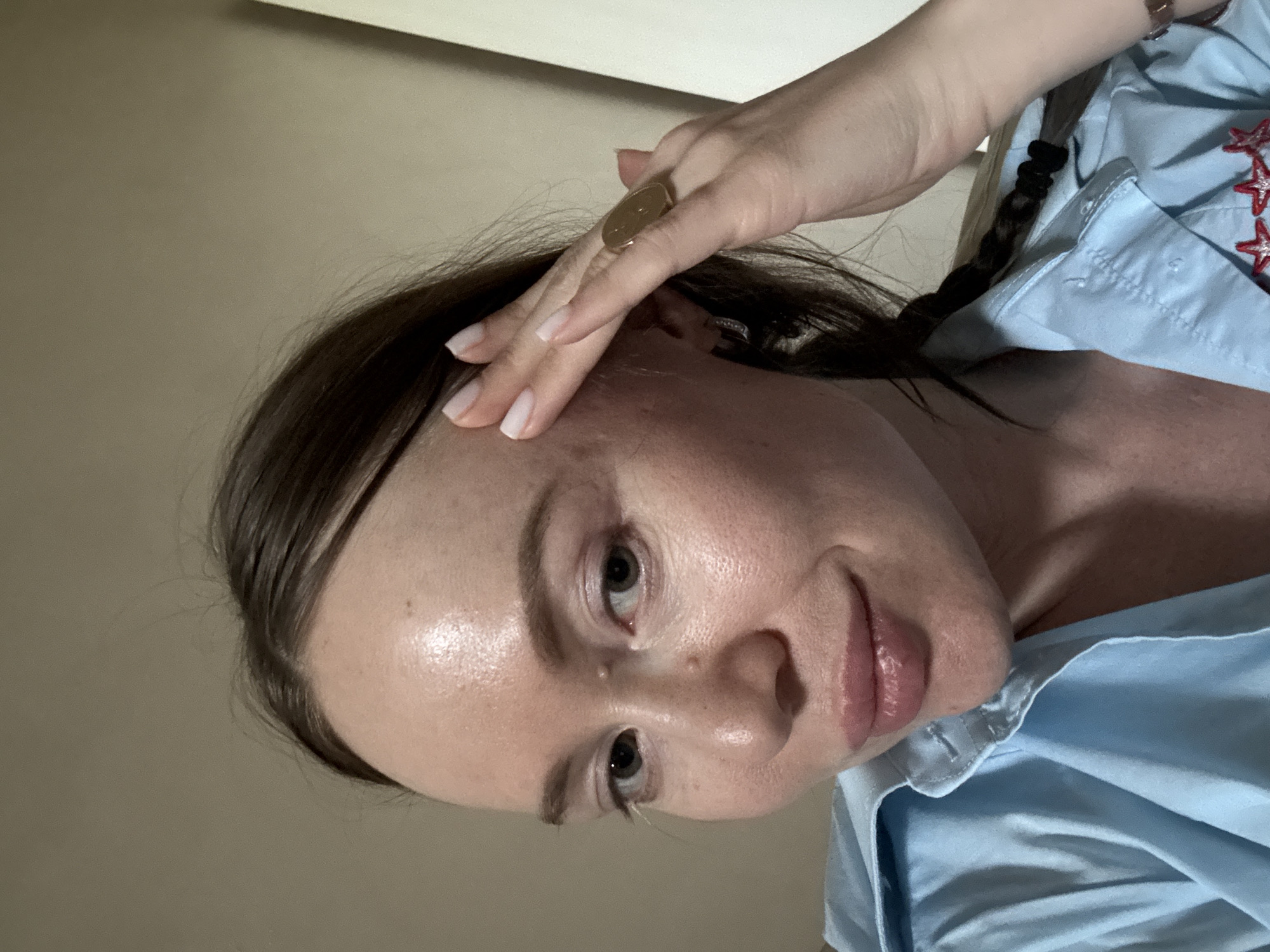 A young Caucasian woman with light skin, brown hair, and light brown eyes is pictured. She is wearing a light blue button-down shirt. Her right hand is raised to her forehead, and she is wearing a large, oval-shaped ring on her right index finger. Her hair is pulled back into a braid. The background is a neutral, plain wall. The lighting suggests an indoor setting, possibly a home environment. No specific details indicate the location in the city of Krasnoyarsk, Russia.