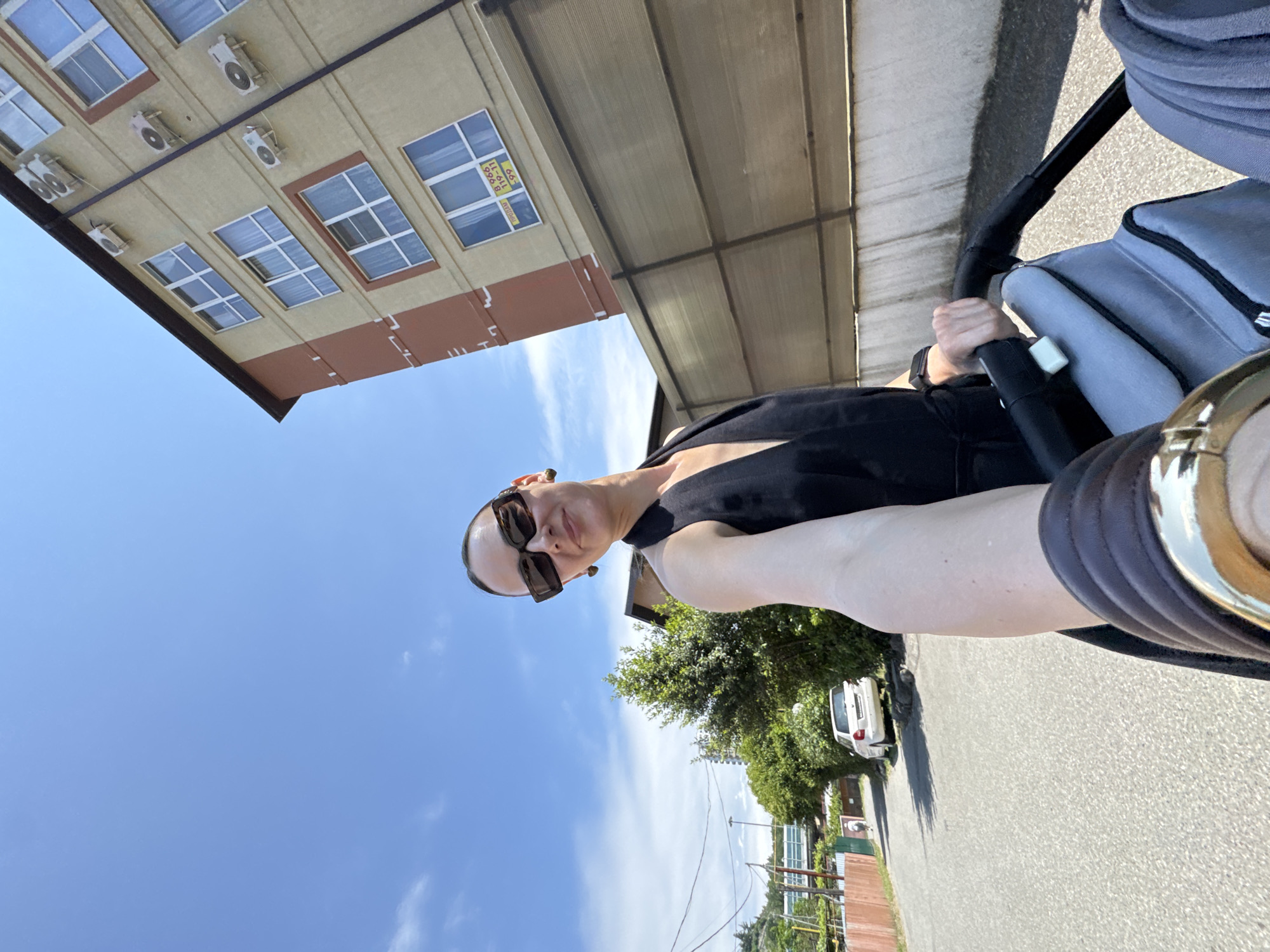 A woman wearing sunglasses and a black dress is pushing a stroller on a street in Sochi, Russia. The woman is in the foreground, holding the stroller handle with her right hand. Behind her, a multi-story building with many windows and air conditioners is visible. The building appears to be under construction or renovation, as some windows are covered with yellow signs. A fence runs along the side of the street, separating it from the building. In the background, there are trees and a white car parked on the side of the road. The sky is blue with some clouds. The street is paved and appears to be a residential area.