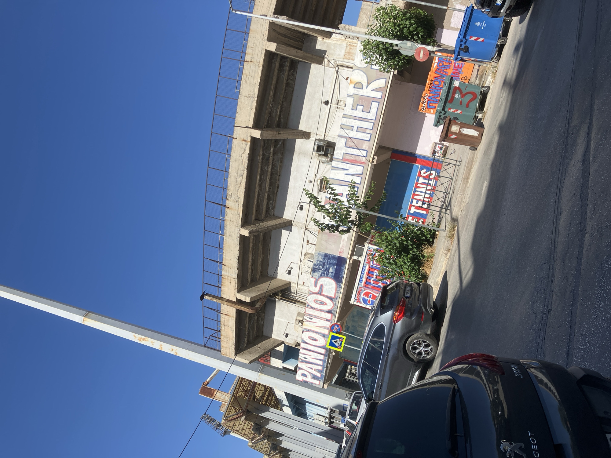 The image shows a street scene in front of a stadium. The stadium has a concrete structure with a rusted metal railing at the top. Large white letters painted on the side of the stadium spell out "PANIONIOS" and "PANTHER". Below the stadium, there are several cars parked on the street, including a dark gray SUV and a dark gray hatchback. There are also several trash bins, including a green bin with a red "3" on it, a brown bin, and a blue bin. Trees and bushes are visible on the right side of the image. The sky is clear and blue.