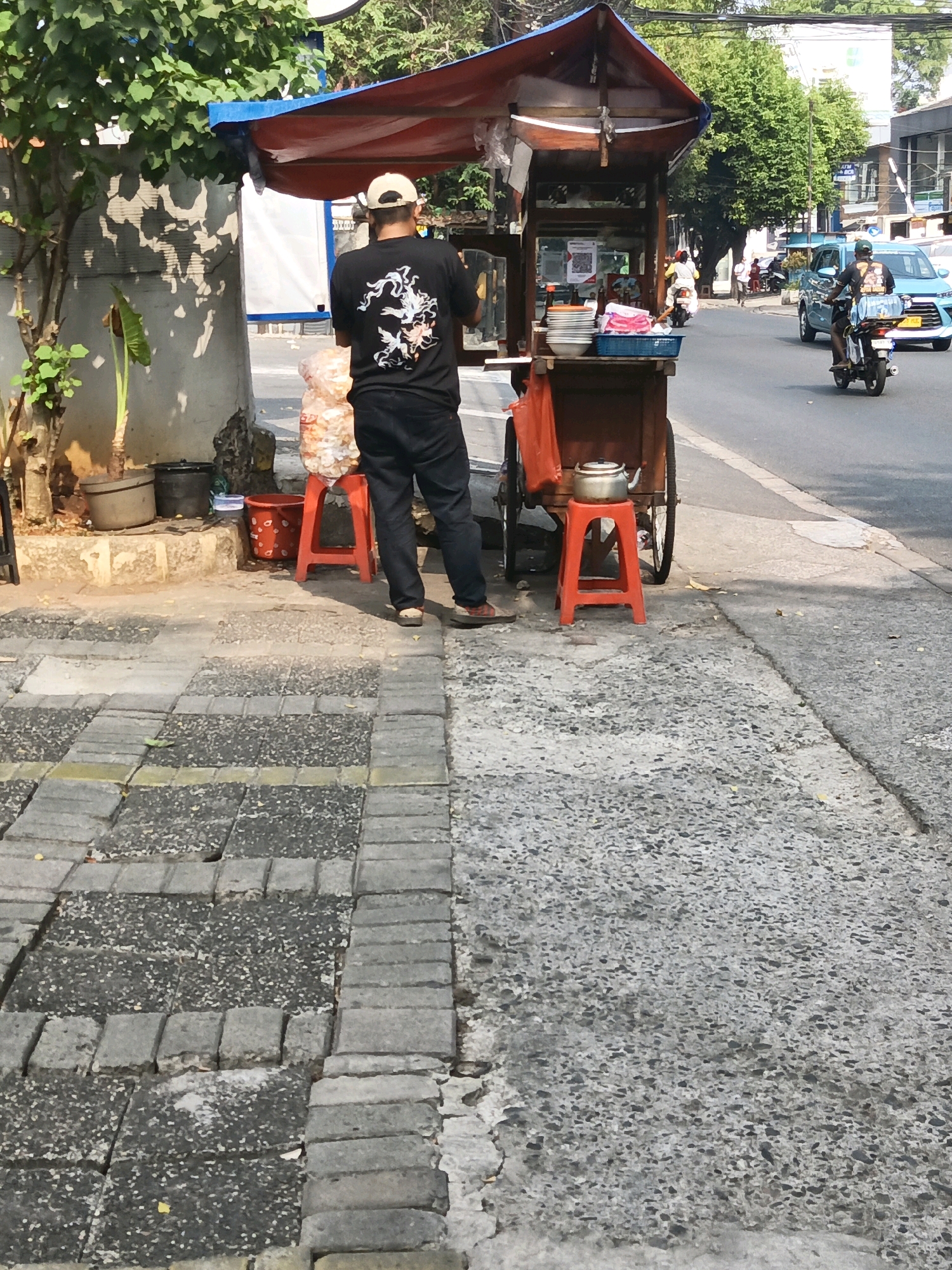A person wearing a black t-shirt with a graphic design and a beige baseball cap stands beside a food cart on a sidewalk in South Jakarta, Indonesia. The food cart has a red and blue awning and appears to be selling snacks. A large bag of chips is on a red stool next to the cart. The sidewalk is made of interlocking concrete pavers. In the background, a blue car drives past and a motorcycle is parked. Trees and buildings line the street.