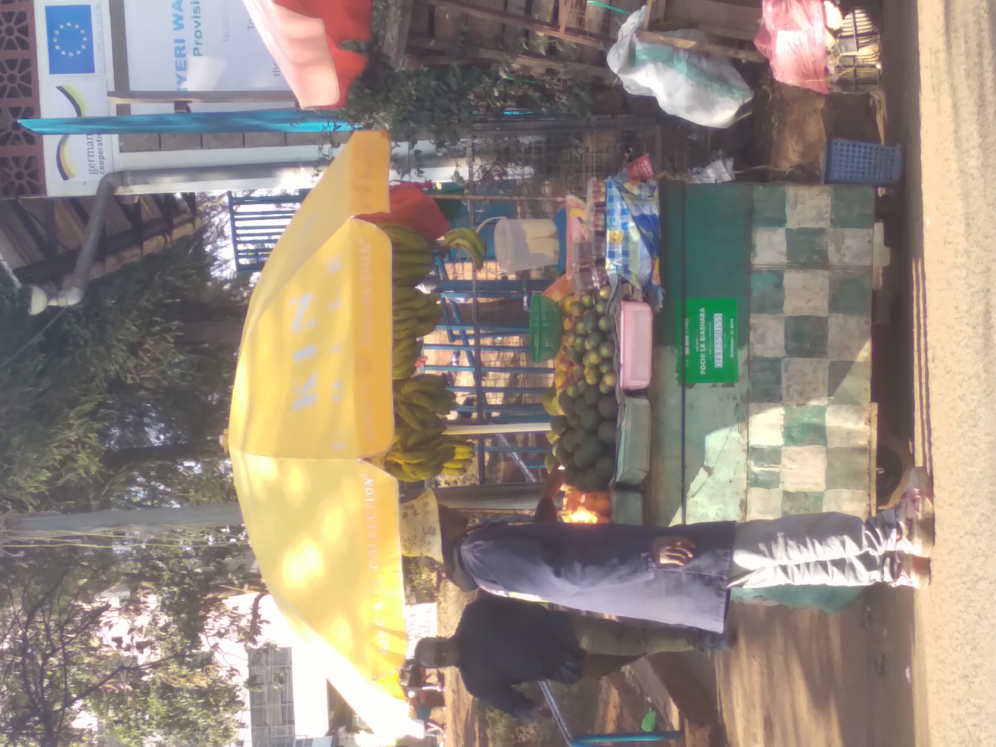 The image depicts a street scene in Nyeri, Kenya, near a fruit stall. A yellow umbrella, possibly a part of the stall's setup, is prominently featured. Several individuals are visible: a person in a dark shirt and trousers walking towards the right, another person wearing a blue coat and light-colored pants interacting with the fruit stall, and other figures in the background. The stall itself displays an assortment of fruits, including bananas, avocados, and limes, arranged in containers. A green and white checkered pattern adorns the lower part of the stall. Various objects, such as plastic bags, a blue crate, and a wooden ladder, are scattered around the stall. Signs are visible in the background, one with German text and the EU flag, and another with text that includes "YERIE WA" and "Provisi". The overall environment suggests a busy outdoor market or roadside selling area.