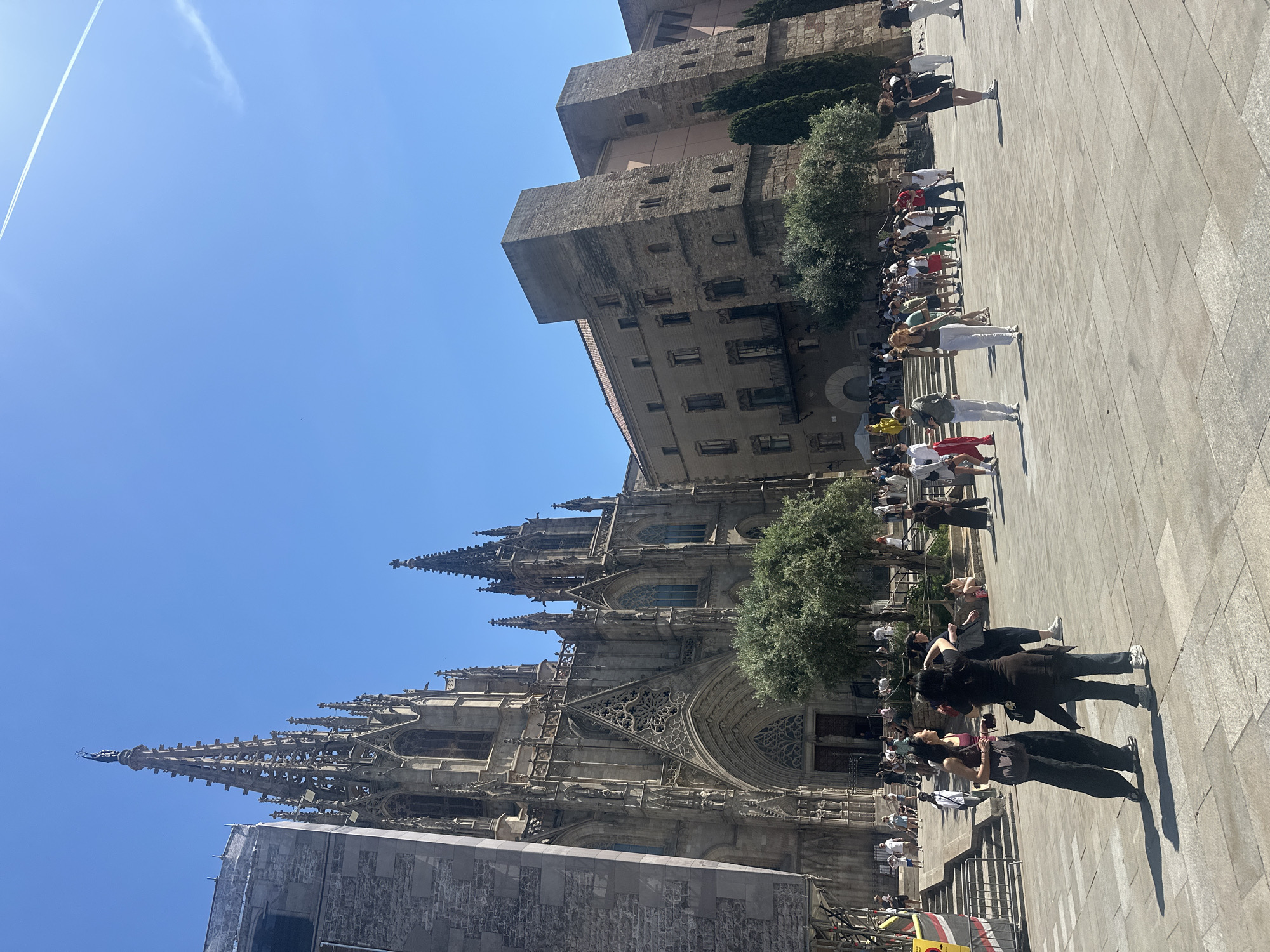 The image shows the Gothic facade of the Barcelona Cathedral, also known as the Cathedral of the Holy Cross and Saint Eulalia, in Barcelona, Spain. The photograph is taken at ground level, looking upwards towards the cathedral's spires and towers. A group of people is standing on a stone-paved square in front of the cathedral. The sky is clear blue. Another stone building with multiple stories is visible to the right of the cathedral. A bright streak from an airplane is visible at the top left of the frame.