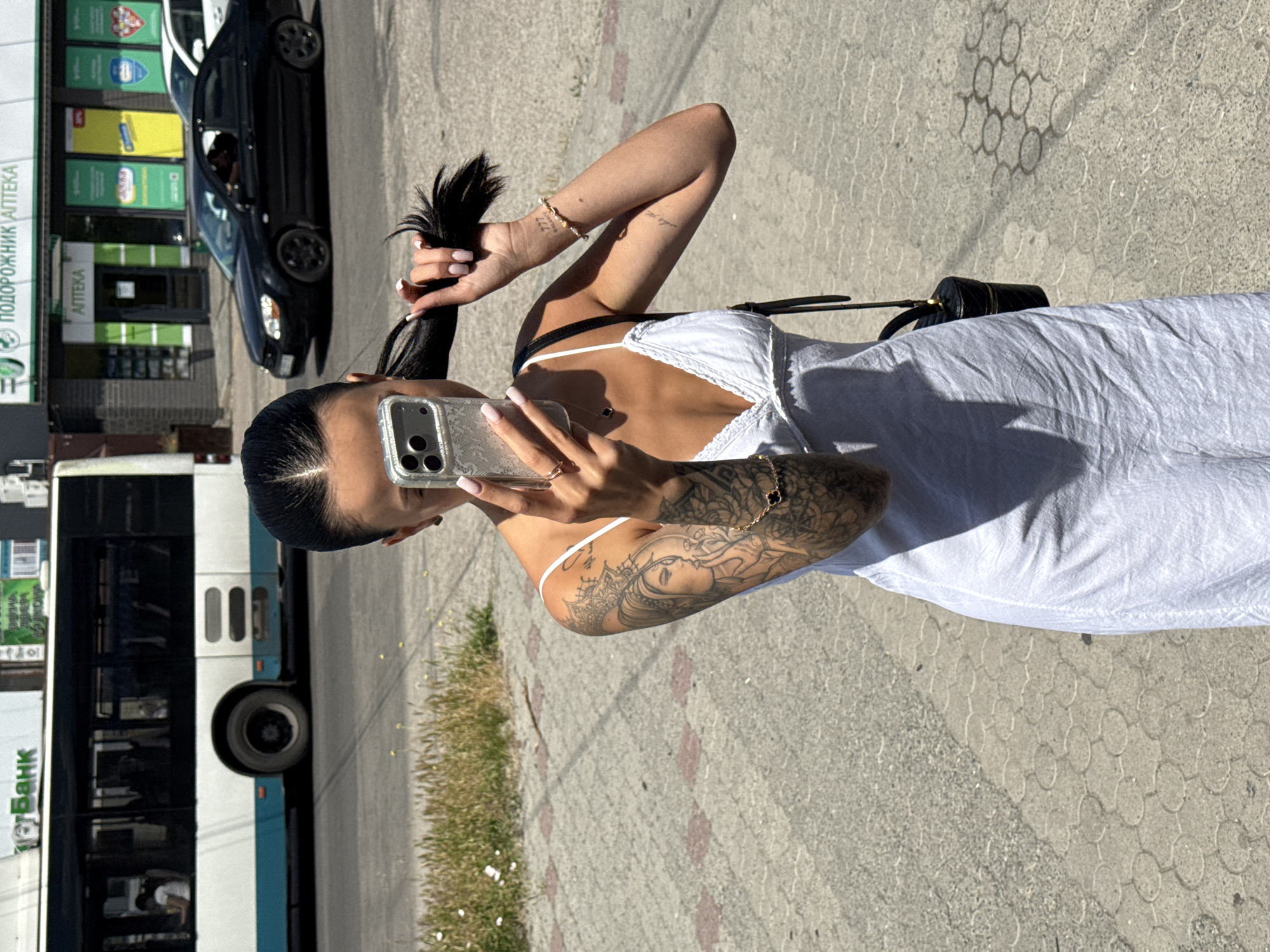 A young woman with dark hair pulled back in a ponytail stands on a paved sidewalk, taking a mirror selfie with her smartphone. She is wearing a white sundress and has tattoos on her left arm and shoulder. She holds her hair in her right hand as she snaps the photo. In the background, a bus is parked on the street, and several cars are visible, along with buildings that appear to be shops. The environment suggests an urban setting on a sunny day.