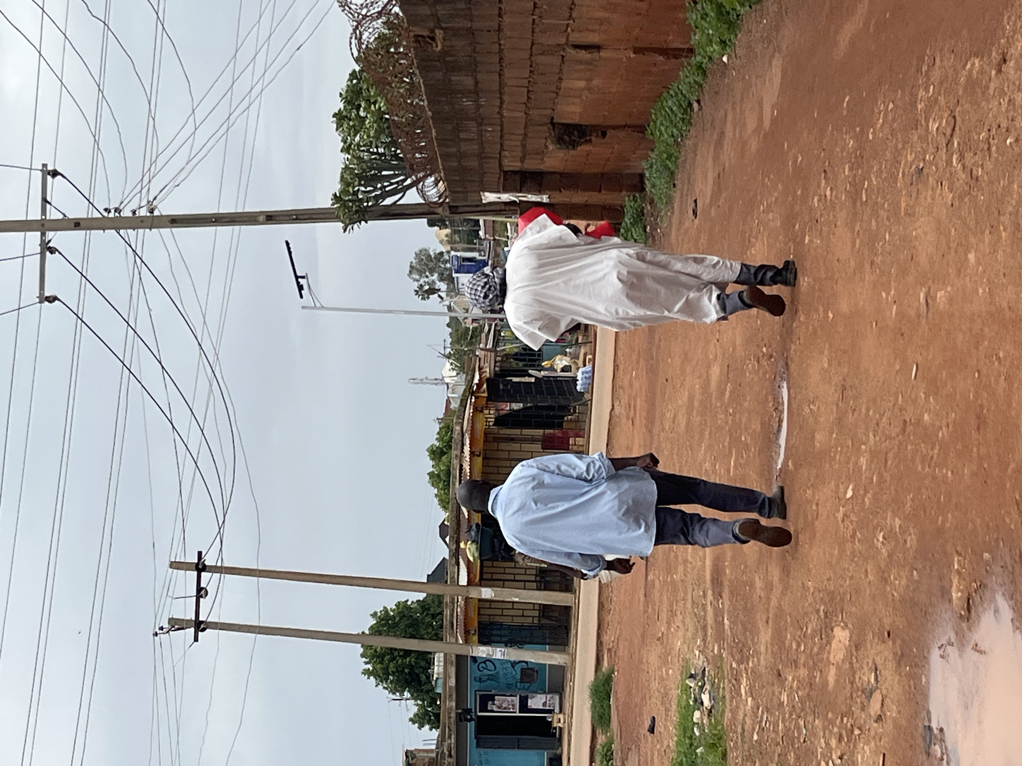 The image shows two men walking down a dirt road. The man in the foreground is wearing a light blue long-sleeved shirt and dark pants. He is walking away from the camera. The man behind him is wearing a white robe with a red headdress. He is also walking away from the camera, and appears to be carrying something red. There are power lines overhead and a brick wall on the left side of the road. The ground is covered in dry, reddish-brown dirt, with some patches of green grass and a puddle of water on the right side. In the background, there are some small buildings with corrugated roofs and trees. The sky is overcast, suggesting it might be daytime or cloudy weather. The overall scene appears to be in a rural or semi-urban area. There is no visible text in the image.