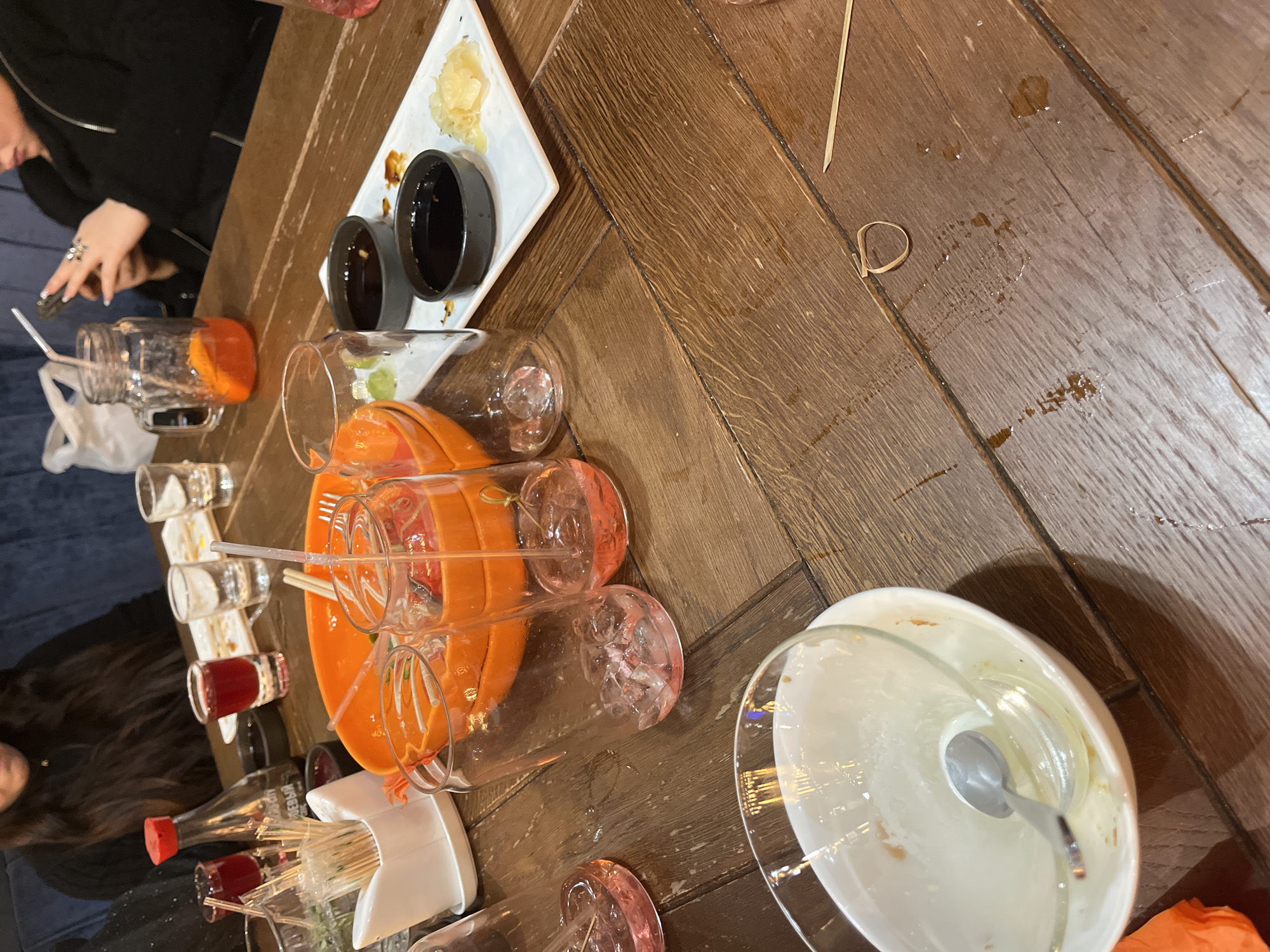 The image captures an overhead view of a wooden table with a distinctive herringbone pattern, strewn with items from what appears to be a social gathering or meal. The table is cluttered with numerous empty or partially filled glasses and bottles. Among these are several shot glasses, some containing residual red liquid, and a glass jar holding orange slices and a straw. A small bottle with a red cap bears Cyrillic text, appearing to read "кедровый" (kedrovyy), indicating a cedar-based product, common in Russia. A white holder filled with many thin straws is also on the table.
On a white square plate, two small black bowls, likely for soy sauce, sit next to a pile of pickled ginger, suggesting a meal involving sushi or Asian cuisine. A large, empty white bowl with a spoon rests on its side. Various spills and drops of liquid are visible on the wooden surface, indicating an active event.
Partial views of two individuals are present: one person wearing a black jacket with a prominent silver ring on their finger, and another with long, dark hair. Both are engaged with items on the table. A small, light-brown paper loop and a thin wooden skewer are also visible on the table.
The scene depicts the relaxed aftermath or ongoing state of an indoor social event, possibly at a restaurant or bar. The indoor setting, absence of natural light, and the type of beverages and food items suggest an evening gathering. The Cyrillic text on the bottle aligns with the given location context of Saint Petersburg, Russia.