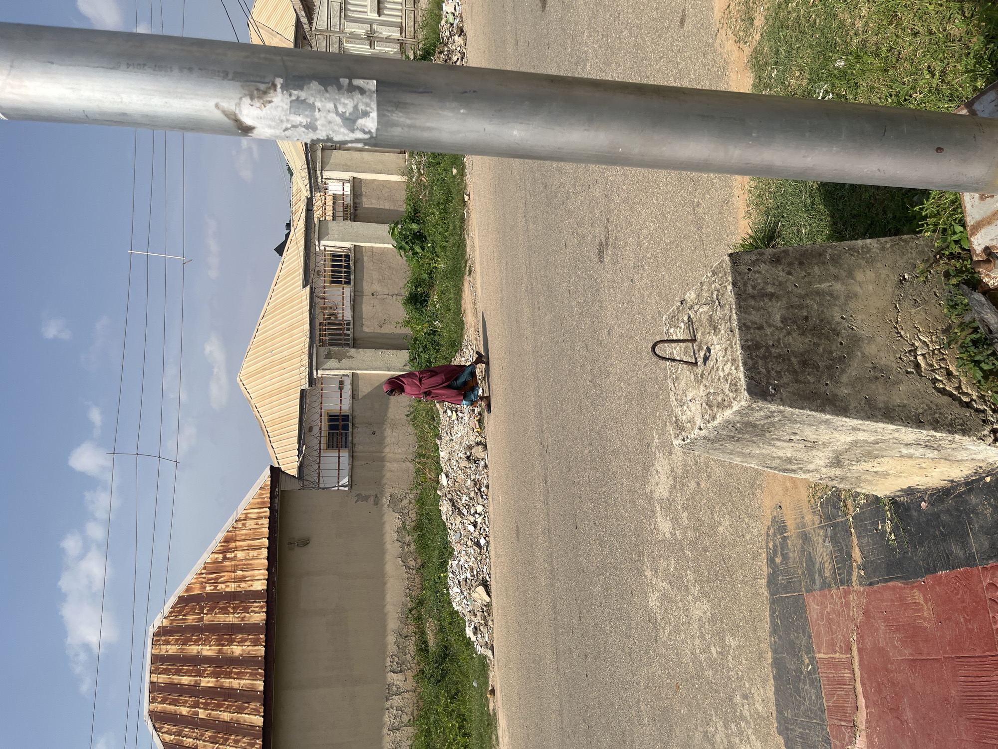 A wide shot captures a street scene in Abuja, Nigeria, under a clear, bright sky with scattered clouds. In the foreground, a grey concrete base with a U-shaped metal hook sits on a patterned sidewalk made of red and black tiles. To the right, a silver metal pole is prominently positioned, obscuring part of the background. The street itself is paved with asphalt.
Across the street, a pile of debris, consisting of rubble and refuse, is visible along the edge of the road. Behind the debris, a building with a light-colored wall and a corrugated, rusty metal roof is situated. Further back, another building with a similar roof style is visible, bordered by a fence with barbed wire. A person, likely a woman, is walking from left to right across the scene, draped in a maroon garment that covers her head and body. She is walking on the asphalt street, to the left of the silver pole. The environment appears to be a residential or commercial area, possibly undergoing construction or experiencing neglect due to the presence of the debris. The overall impression is one of everyday life in a Nigerian urban setting.
