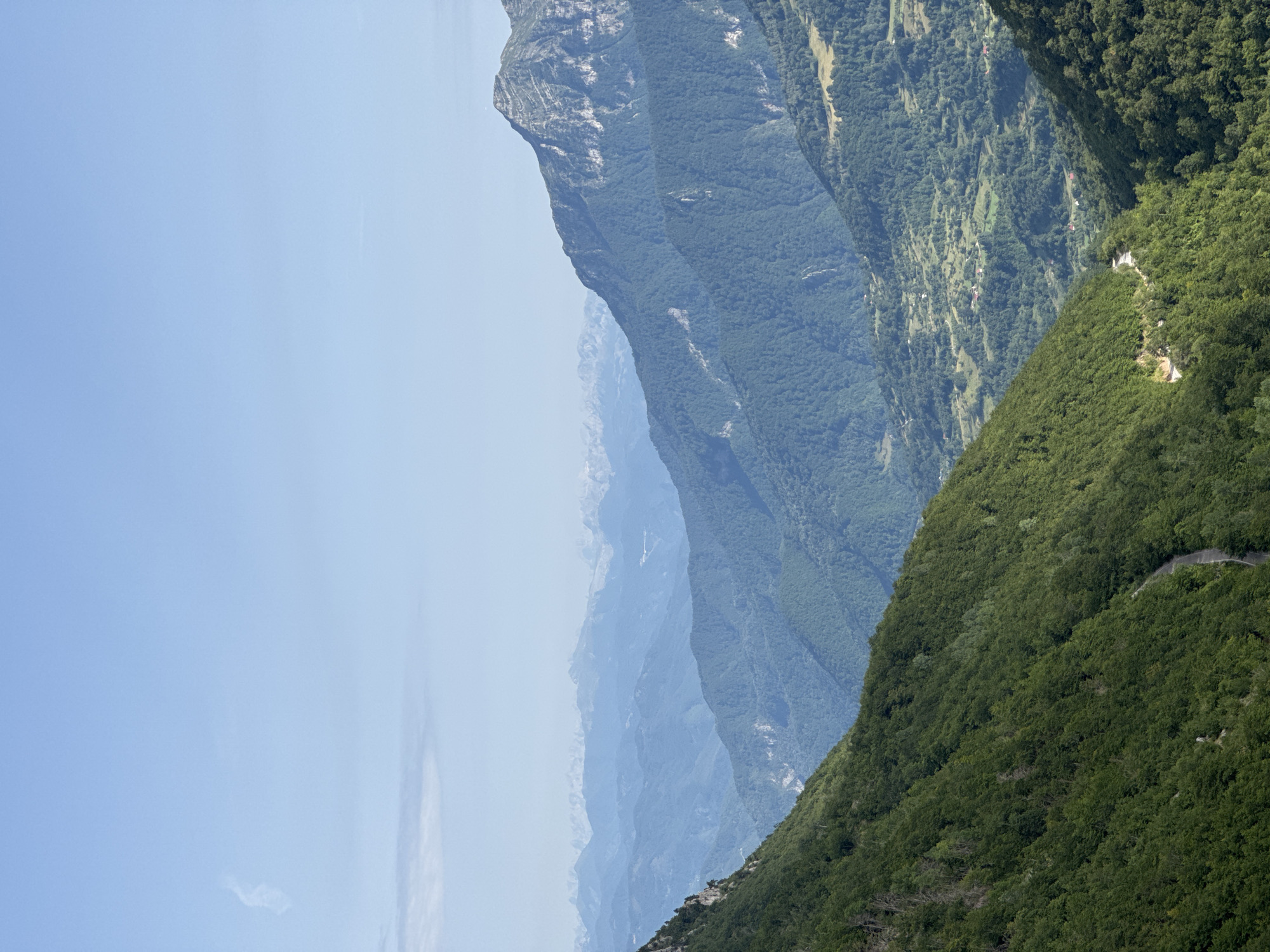 The image depicts a vast, mountainous landscape under a clear blue sky with a few wispy clouds. The foreground is dominated by a steep, densely forested mountainside on the left, with a winding road visible. The middle ground features rolling, tree-covered hills and a valley with scattered houses and agricultural fields. In the background, a series of progressively lighter blue mountain ranges recede into the distance, suggesting a significant elevation and a hazy atmosphere. The scene is devoid of people or specific man-made structures, emphasizing the natural, rugged environment. The lighting suggests it is daytime, likely mid-morning or afternoon, with the sun casting light and shadows that highlight the contours of the terrain. The overall impression is one of natural grandeur and a remote, untouched wilderness.