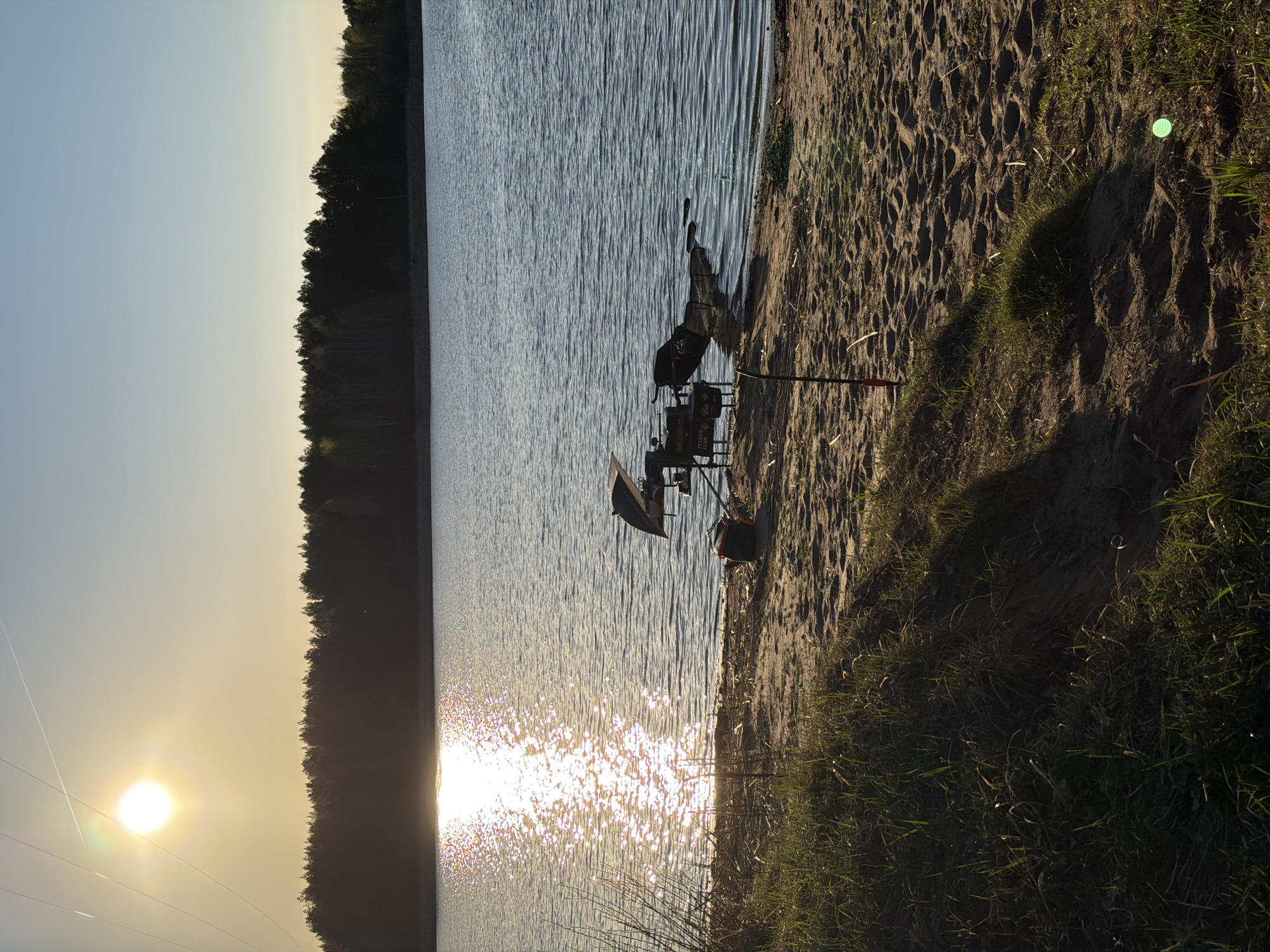 The image shows a sandy beach on a body of water, possibly a lake or a wide river, under bright sunlight. A fishing setup is on the beach, including a rod stand, tackle boxes, and a bucket. A fishing rod is planted in the sand. The water reflects the sunlight, creating bright spots. In the background, there's a line of trees, indicating a forest or wooded area across the water. The scene suggests an early morning or late afternoon, based on the sun's position and the resulting shadows. The specific location in Chaykovsky, Russia, is difficult to determine without more context, but the image portrays a common lakeside fishing scene. There are no people visible in the image.