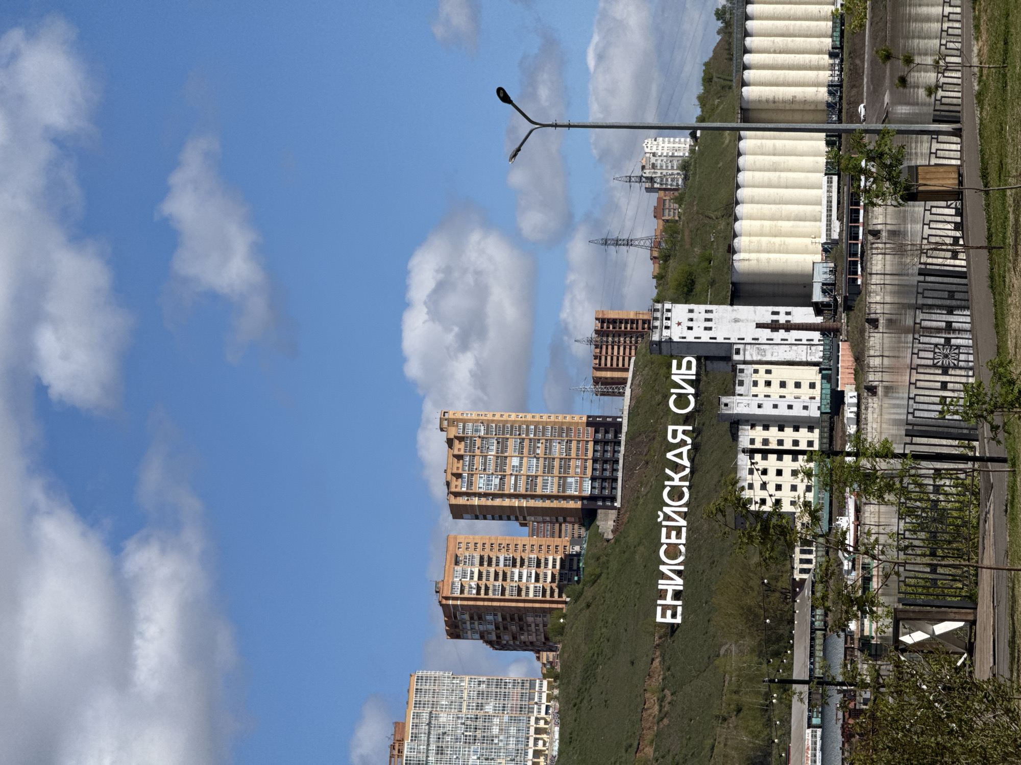 The image shows a hillside in Krasnoyarsk, Russia, with residential buildings on top of the hill and the words "ЕНИСЕЙСКАЯ СИБ" (Yeniseyskaya Sib) written on the hillside. In the background, there are large white cylindrical structures, possibly storage containers. In the foreground, there's a fence, a street lamp, and a few trees. The sky is blue with scattered clouds. Electrical power lines can also be observed.
