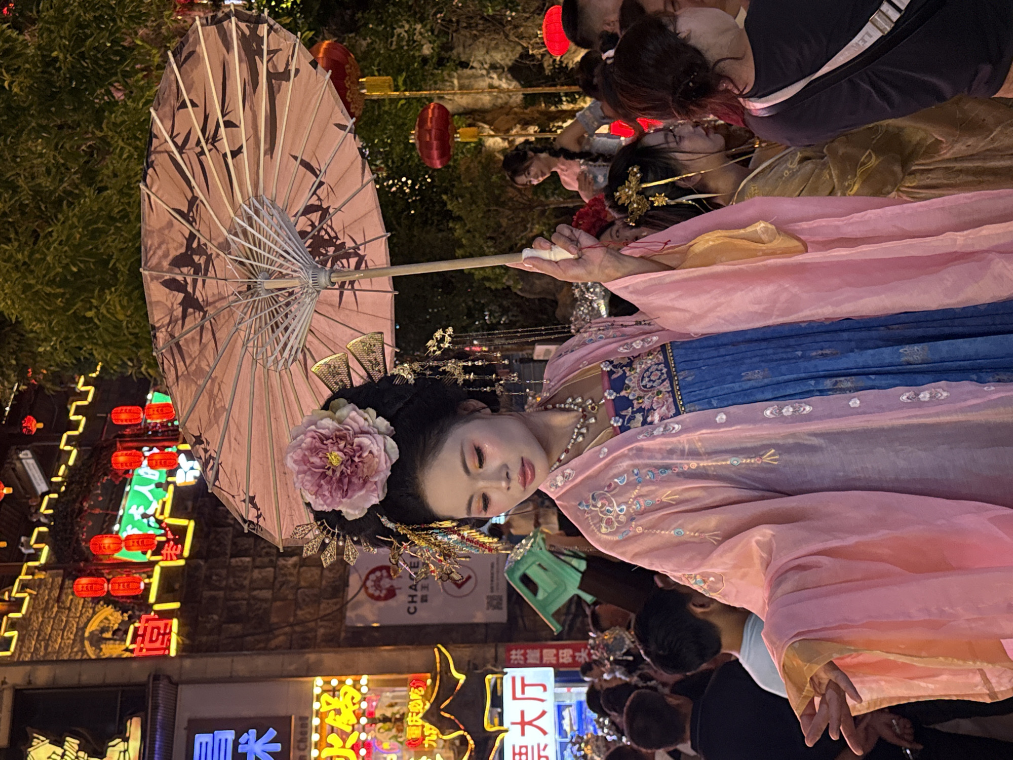A young East Asian woman, adorned in traditional Chinese attire including a pink silk robe with embroidered details and a blue pleated skirt, stands at the forefront of the image. She holds a pink paper umbrella patterned with bamboo leaves over her head and wears an elaborate headdress topped with a large pink flower. Her makeup is pronounced, with colorful eyeshadow and rosy cheeks. Her left hand is extended forward in a graceful gesture. Behind her, a crowd of people mill about a bustling street scene at night. Neon signs with Chinese characters illuminate the background, suggesting a commercial or entertainment district. Red lanterns hang overhead and from trees, adding to the festive atmosphere. The overall scene depicts a cultural performance or event in Yuzhong District, China, likely during an evening festival or celebration.