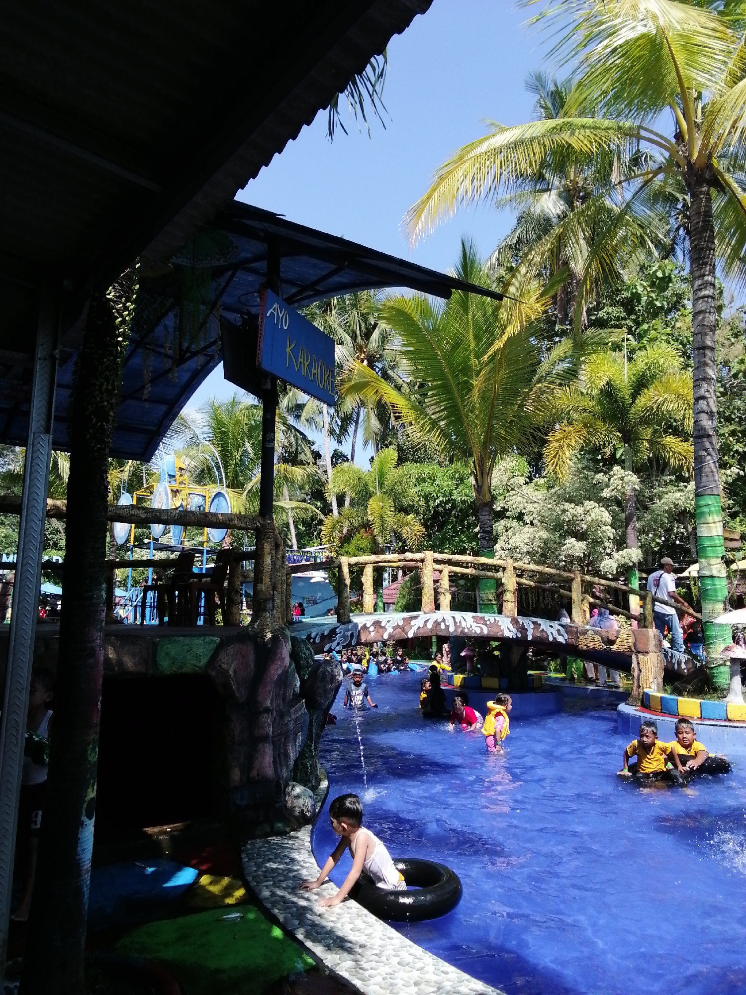 The image depicts a lively scene at a water park, likely located in Tegalwangi, Indonesia, suggested by the lush tropical vegetation and palm trees. In the foreground, a young boy in a white tank top and dark shorts is sitting on the edge of a bright blue pool, reaching towards a black inner tube in the water. The pool is filled with people, many of whom are children wearing colorful swimwear and floatation devices, suggesting active recreation.
To the right, two young boys in yellow shirts are sitting on black inner tubes, seemingly enjoying the water. In the middle ground, a wooden bridge with a decorative stone railing spans the pool. Several individuals, including adults and children, are visible on and around the bridge. Further in the background, a water slide structure with blue elements can be seen.
Above the pool area, a covered structure with a corrugated roof partially shades the scene. A blue sign with white text that reads "AYO KARAOKE" is prominently displayed. The overall environment is one of a recreational facility with a vibrant, sunny atmosphere. The water is a clear, inviting blue, and the surrounding greenery provides a natural, tropical backdrop. The presence of a karaoke sign hints at the entertainment offerings of the water park.