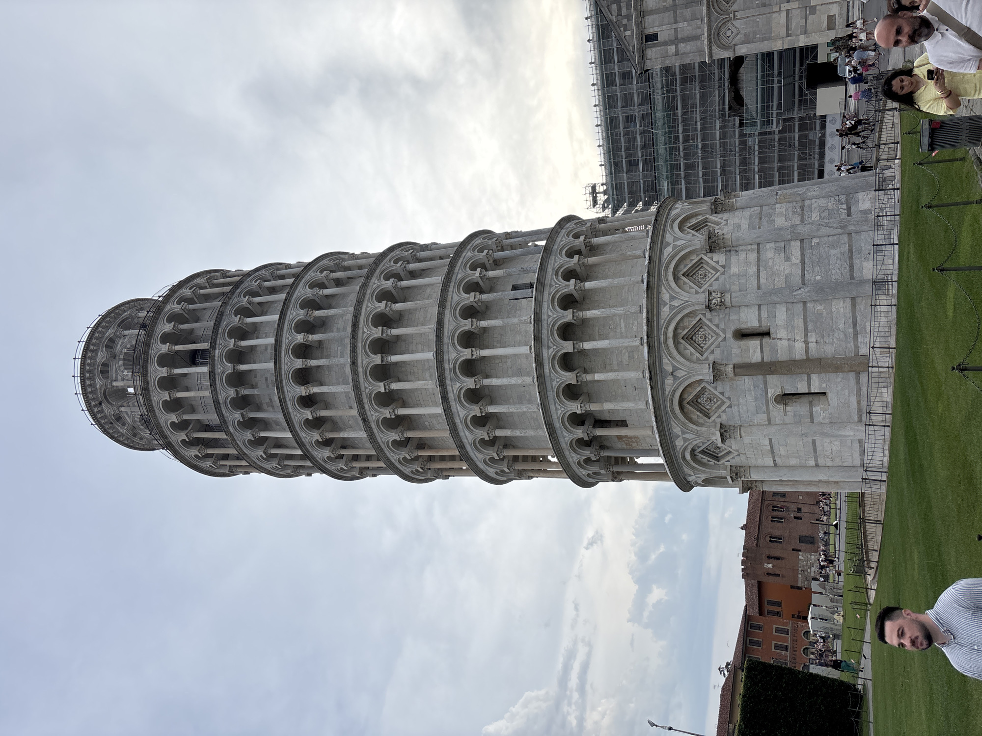 The Leaning Tower of Pisa dominates the scene, positioned slightly off-center and tilted noticeably to the right. The tower's multiple tiers of arched colonnades are prominently displayed. Scaffolding is visible on a structure adjacent to the tower. The foreground includes a grassy area with a metal chain fence. People are present, some appearing to take pictures. In the background, buildings with red tile roofs are partially visible. The sky is overcast with a pale, diffused light.