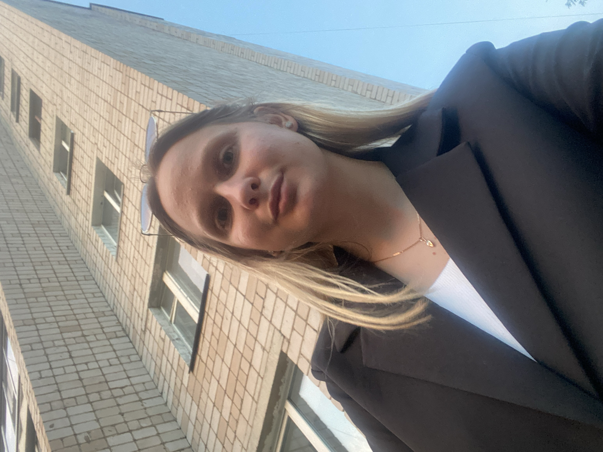 A young woman with blonde hair and fair skin is captured in a low-angle selfie, standing in front of a multi-story brick building. She is wearing a black blazer over a white top and a delicate necklace. Sunglasses are perched on her head. The building, characteristic of Soviet-era architecture, features rows of identical windows and is constructed from light-colored bricks. The sky above is a clear blue. This scene is likely located in the city of Chaikovsky, Russia, a city known for its industrial development and associated residential buildings.