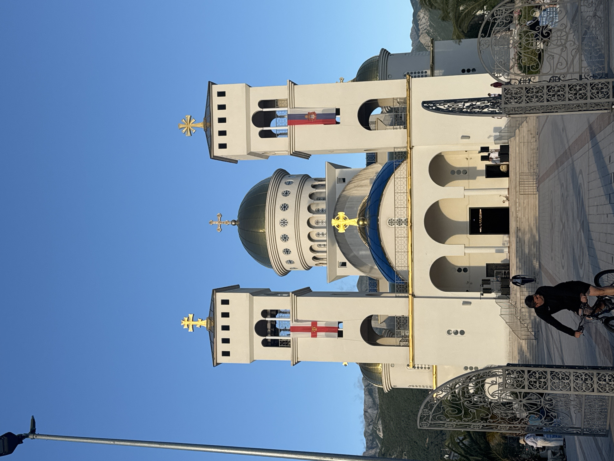 The image depicts the elaborate Cathedral of Saint John Vladimir in Bar, Montenegro, under a clear blue sky. The grand, white Orthodox church dominates the scene, featuring multiple domes; a large central dome with a greenish-gold top and decorative elements, and another with a blue and white patterned surface. Two prominent bell towers flank the central structure, each topped with a golden, ornate cross.
Flags are displayed from the bell towers. On the left tower, the Serbian national flag (red, blue, white tricolor with a coat of arms) hangs. On the right tower, the flag of the Serbian Orthodox Church is visible, featuring a red cross on a white field, flanked by four stylized Cyrillic 'C's (standing for "Samo Sloga Srbina Spasava" - Only Unity Saves the Serbs). The church facade is adorned with arched windows, golden detailing, and intricate patterns.
In the foreground, a person wearing dark clothing rides a bicycle on a paved area to the right. An ornate white metal gate with scrollwork details runs across the lower-middle section of the image, partially obscuring the church's lower levels and steps. Other indistinct figures are visible near the church entrance and behind the gate. A street lamp post is in the bottom left. The background features green foliage, including palm trees, and distant mountains.
The scene is brightly lit by natural sunlight, suggesting either morning or late afternoon, with no clouds in the sky. The overall impression is one of architectural grandeur and peaceful outdoor activity.