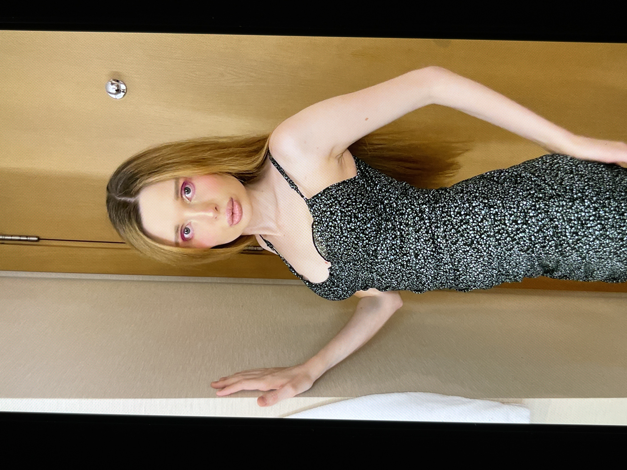 This media file shows a young woman with blonde hair and pink eye makeup, wearing a black and white floral print dress. She is posing with her arms extended, one resting on a light-colored surface and the other raised. The background appears to be a room with a tan wall and a light-colored countertop or shelf. There is a round, reflective object on the wall behind her, possibly a doorknob or a piece of decor. The lighting suggests an indoor setting, possibly during the day. The woman's expression is neutral. There is no visible text in the image.
