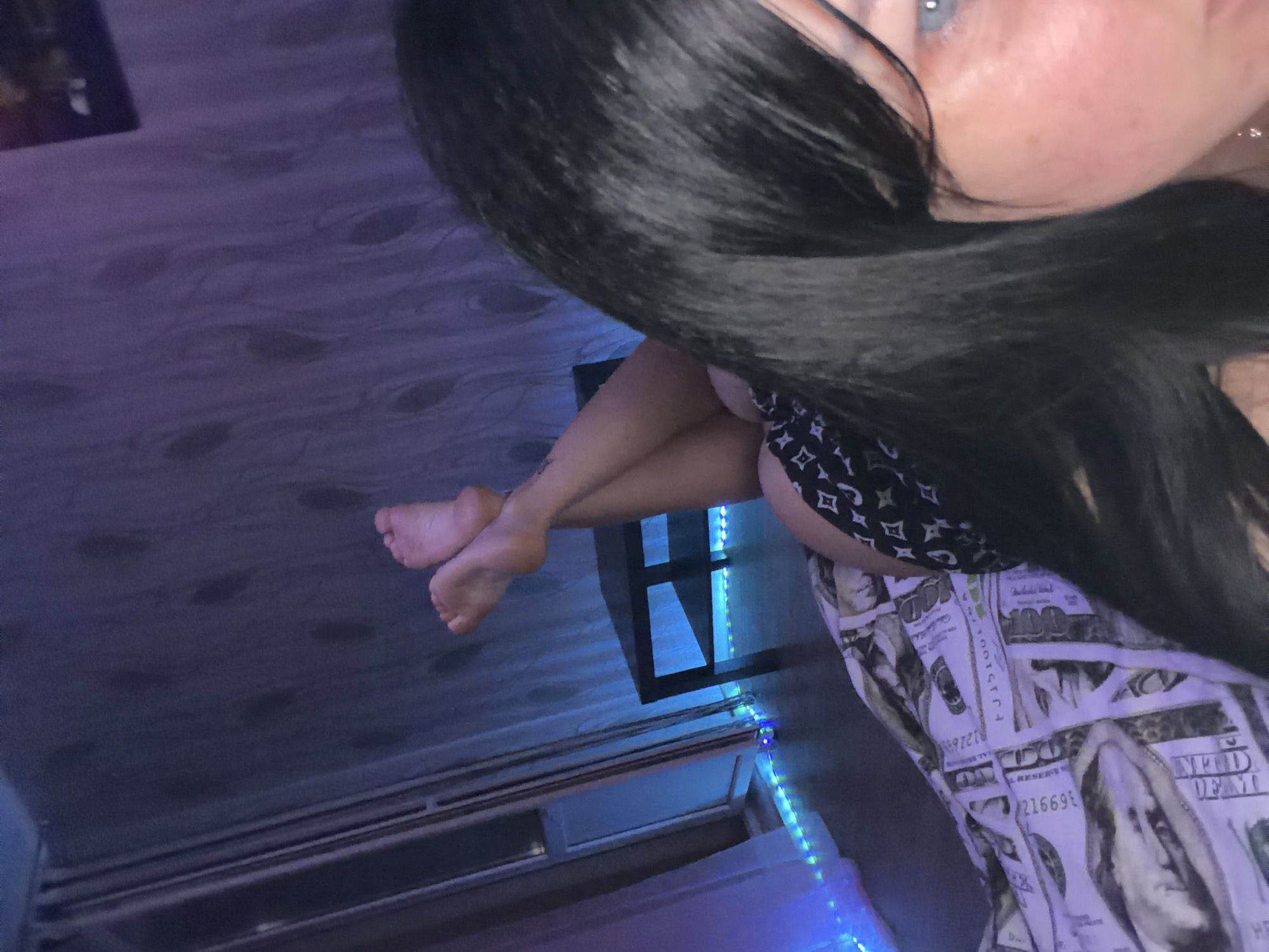 This image shows a person lying down, with their legs crossed and feet in the air. The person has long, dark hair and is wearing patterned shorts. They are lying on a blanket with a money print. In the background, there is a dark table and a wall with a patterned wallpaper. There are also blue LED lights in the scene, suggesting it is nighttime or a dimly lit environment. The location is identified as Ufa, Russia.