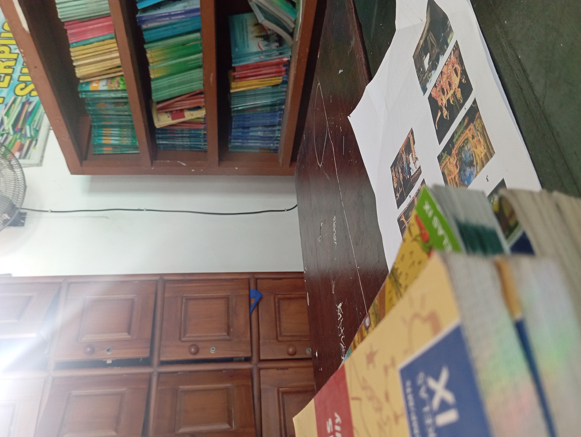 The image depicts a cluttered wooden desk in a room that appears to be a library or classroom. A stack of books, one with "KELAS IX" visible, lies on the left side of the desk. Several printed images are scattered across a sheet of paper on the right side of the desk. In the background, a wooden cabinet with multiple doors is visible on the left, and a bookshelf filled with colorful books is on the right. A ceiling fan is partially visible at the top center. The lighting suggests the presence of sunlight streaming in from the left, creating a lens flare effect. The overall impression is one of a functional, perhaps slightly disorganized, educational or study space. There is no indication of the specific location within Petungasri, Indonesia, beyond the general setting of a room with books and study materials.