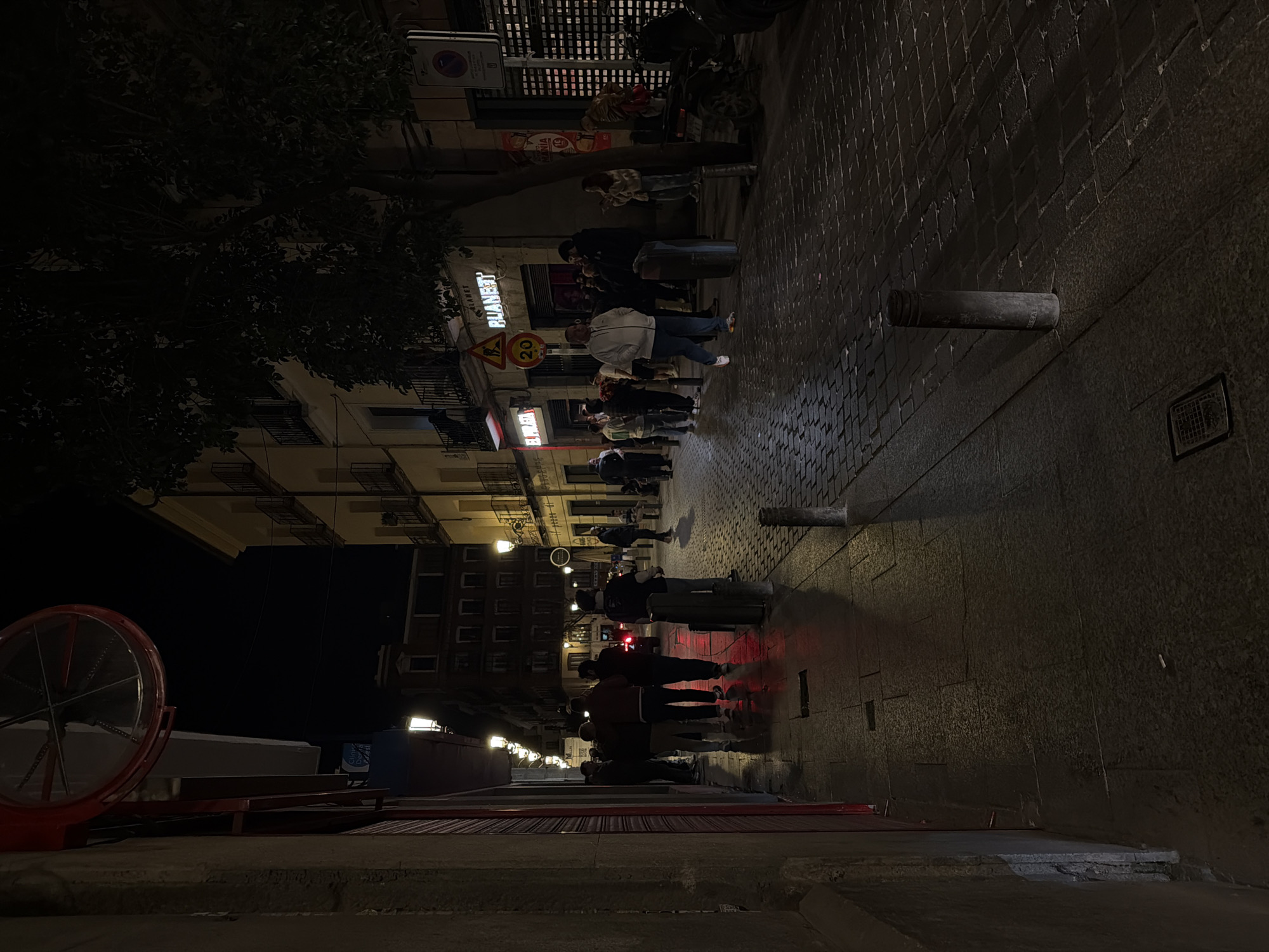 The image depicts a nighttime street scene on Calle de la Luna in Madrid, Spain. The narrow street is paved with dark, reflective cobblestones, bordered by concrete sidewalks. Multi-story buildings, characteristic of Madrid's architecture with wrought-iron balconies, line both sides of the street. The dark sky is illuminated by artificial light from numerous lantern-style streetlights and commercial signs, casting elongated shadows.
On the left, a building facade, partially red-painted at the lower level, features a large, red-framed circular convex mirror mounted high. On the right, commercial establishments are visible, including a business with a prominent red neon sign reading "EL PIRATA" and another further down with a white-lettered sign for "PLANET." A large tree with dark foliage stands on the right sidewalk, partially obscuring a building.
Approximately 20 to 30 individuals, predominantly young adults, are present. They are dressed in casual attire, including jackets, hoodies, and jeans. People are observed standing in small groups, conversing, and walking along the sidewalks and in the street. Several dark bollards are positioned along the street and sidewalk. A "PROHIBIDO APARCAR" (No Parking) sign and a yellow triangular warning sign displaying "20" are visible on the right side. A motorcycle is parked on the far right sidewalk, and the red taillights of a distant vehicle are visible further down the street. The scene indicates a lively urban evening environment.
