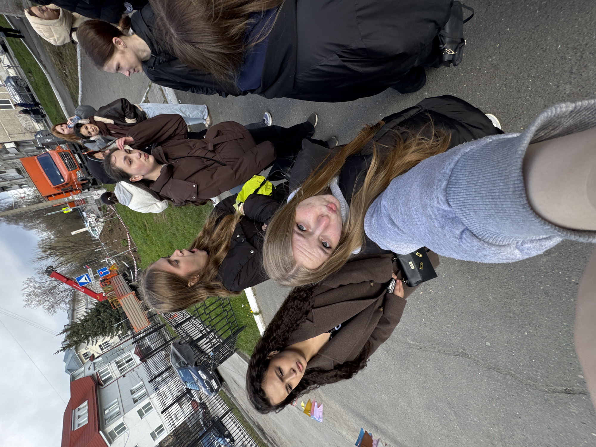 The image depicts a group of at least seven young women gathered outdoors on an urban street or sidewalk. The photo appears to be a selfie taken from a low angle, with the person holding the camera having long blonde hair and looking directly at the viewer with a neutral expression. Another woman with long, dark curly hair is positioned to her right, also looking towards the camera. Other women in the group are seen behind and to the sides, some looking towards the camera, and others looking away or down. They are dressed in casual cool-weather attire, including puffer jackets, longer coats, and hoodies in shades of black, brown, and grey. One woman holds a small black purse with a gold clasp.
The setting is an urban streetscape with an asphalt road and an adjacent sidewalk bordered by grass and a black metal fence. In the mid-ground, a bright orange utility or construction truck with a red crane arm is prominent on the street, suggesting ongoing work or a worksite. Further back, multi-story buildings, typical of residential or mixed-use architecture, line the street. Signs of roadwork or construction are visible, including orange and white striped barriers or tape and a blue road sign with white symbols, though no text is legible. A white car is also parked in the background.
The time of day is daytime, with an overcast sky providing soft, diffused lighting and indicating cool weather. The overall activity appears to be a casual gathering or waiting by the group of women. The general appearance of the architecture and vehicle types are consistent with an urban environment in Perm, Russia.