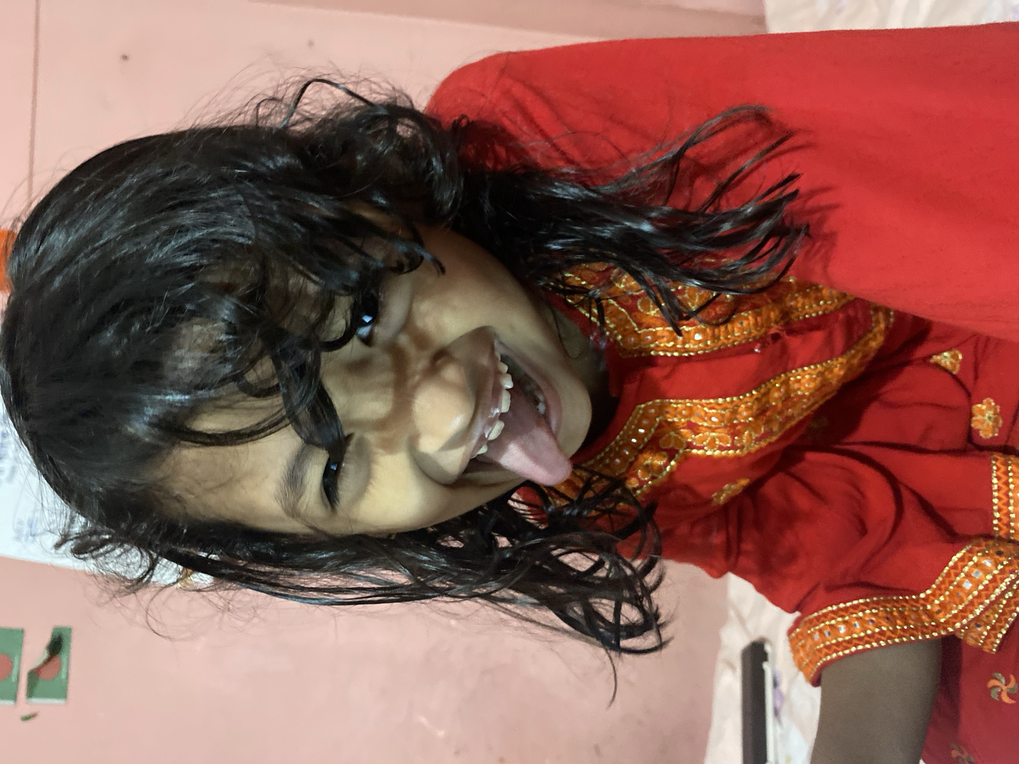 A close-up, eye-level, medium shot captures a young girl with long, dark, wavy hair. She is wearing a bright red garment with intricate gold and orange embroidery around the neckline and sleeves. The girl is looking directly at the camera with a wide smile, her tongue extended outwards. Her teeth, some of which appear to have braces, are visible. The background is a soft pink wall with a framed picture in the upper left corner, featuring two green shapes with red accents. The lighting creates a shadow on the girl's face, emphasizing her playful expression. The scene is likely in a domestic setting, potentially a bedroom, in Dhaka, Bangladesh.