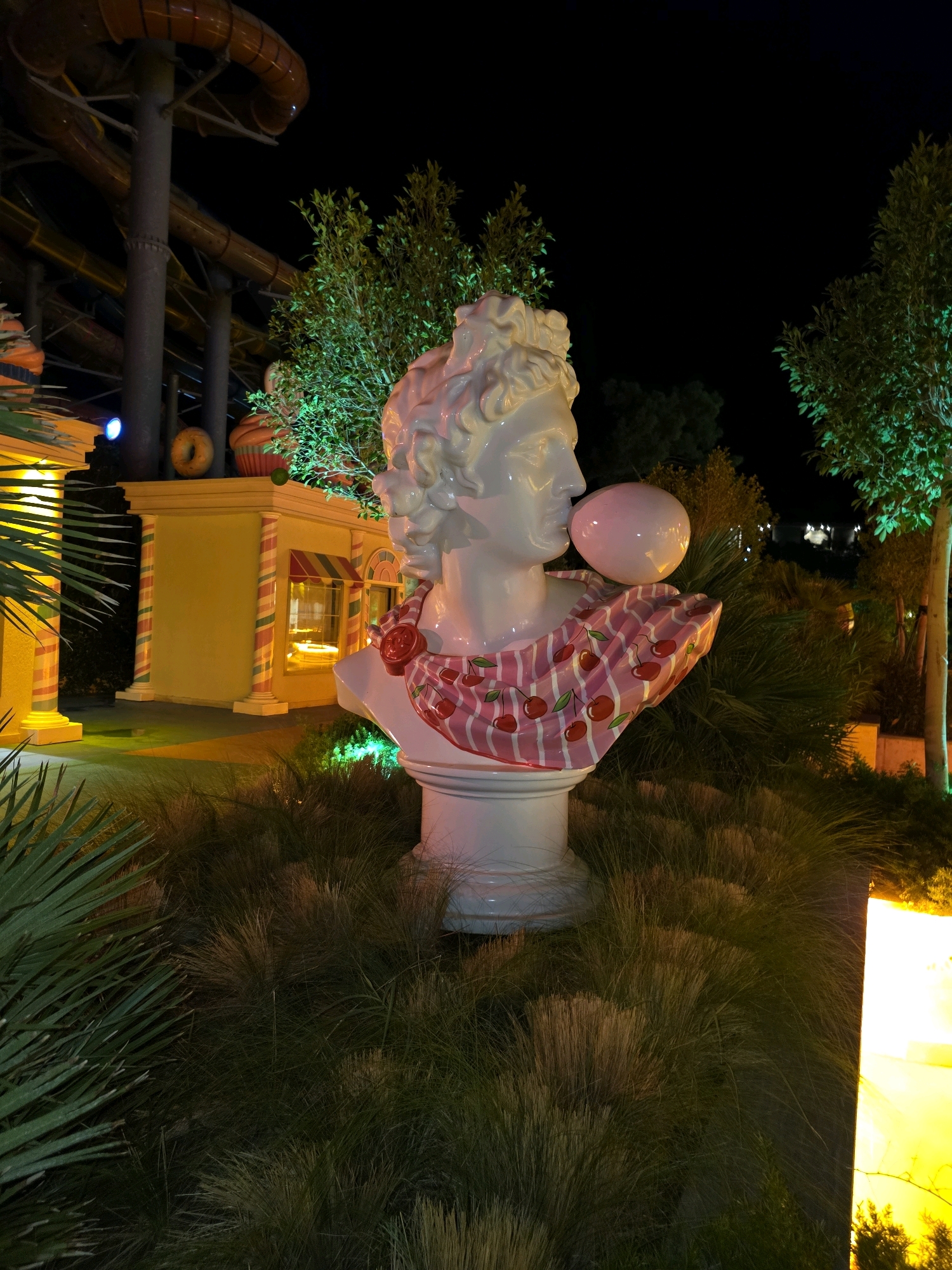 The image depicts a nighttime scene at an outdoor amusement or entertainment venue, possibly a theme park or water park, in Bodrum, Turkey. The central subject is a white bust of a classical figure, possibly a statue of Apollo or a similar deity, depicted blowing a large pink bubblegum bubble. The bust is adorned with a pink and white striped fabric draped around its neck, decorated with red cherries. It rests on a white pedestal, surrounded by tall, wispy grass and palm fronds.
In the background, to the left, are sections of a large, orange water slide structure, illuminated by artificial lights, suggesting a water park environment. Adjacent to the water slide is a brightly lit yellow building with candy-striped pillars, resembling a shop or food stall, possibly themed around sweets or treats. A large donut and a cupcake decoration are visible on top of this building. To the right of the bust, trees are illuminated with green light, creating an atmospheric nighttime setting. The overall scene is illuminated by a combination of spotlights, ambient lighting, and the glow from the structures. No people are visible in the frame. The image captures a whimsical and vibrant atmosphere, blending classical art with a playful, modern aesthetic.