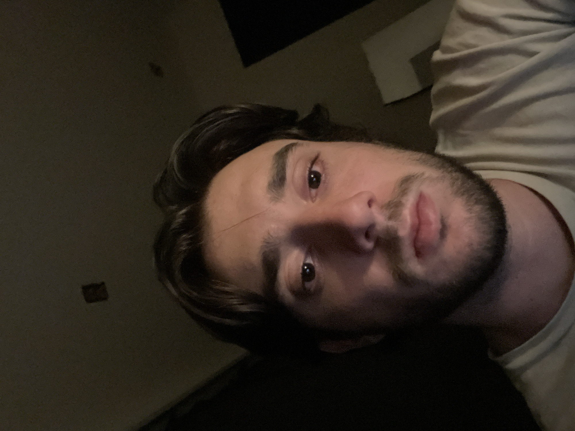 A close-up, low-angle, self-portrait of a young man with dark hair and a beard is shown in a dimly lit room. The man is wearing a light-colored t-shirt. The background is dark, with what appears to be a ceiling visible at the top of the image. There are two small, dark, indistinct objects on the ceiling. A lighter, geometric shape is visible in the background on the right. The lighting suggests the photograph was taken indoors, possibly at night, with the subject illuminated by a nearby light source. The image does not provide any specific details about the location within Huixquilucan de Degollado, Mexico.