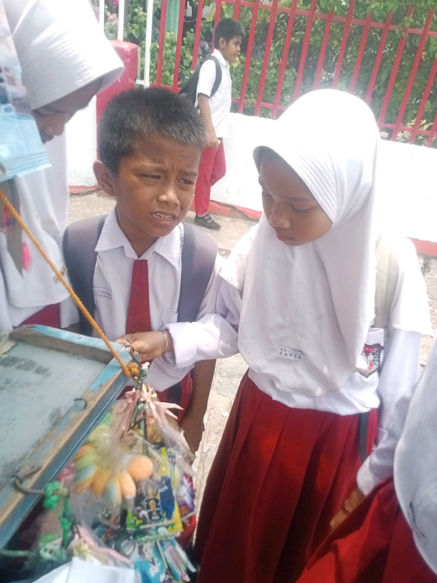 The image depicts a group of schoolchildren in uniform standing near a red fence and a wall. In the foreground, a boy wearing a gray blazer, white shirt, and red tie looks towards a cart filled with various items. Beside him, a girl in a white hijab and a white shirt with a red pleated skirt also looks towards the cart. The cart appears to be a mobile vendor's display, stocked with colorful items, some of which are individually packaged. The children are likely at a school or a public area within the city of Pekanbaru, Indonesia, given the context of schoolchildren in uniform. The background shows more children, some with backpacks, and lush greenery behind the fence. The lighting suggests it is daytime, with sunlight casting shadows. The ground appears to be paved.