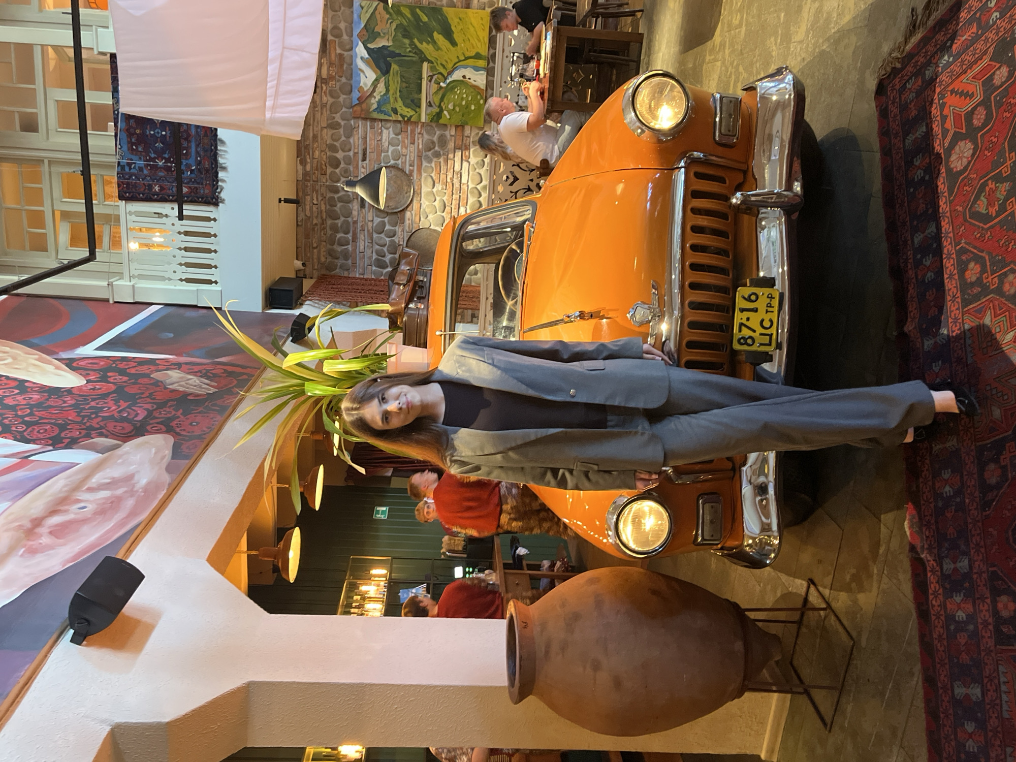 The image captures an indoor scene, possibly a restaurant or a decorative space, featuring a young woman dressed in a grey suit, standing in front of an orange vintage car. The car's headlight is on, and a yellow license plate with the text "87-16 ЦС ТР-Р" is visible. The woman is looking towards the camera with a neutral expression. Behind the car and the woman, other people are partially visible, engaged in activities typical of a restaurant setting, such as sitting at tables and eating.
The background displays elements of interior decor, including stone walls, patterned rugs, and artwork on the walls. Lighting fixtures are also visible, suggesting an indoor evening or dimly lit environment. The overall atmosphere appears relaxed and casual. The scene is filled with objects that create a unique and eclectic ambiance, from the car itself to the large terracotta pot in the foreground and the intricate patterns of the rugs. The image is oriented vertically, with the top of the frame appearing on the left.